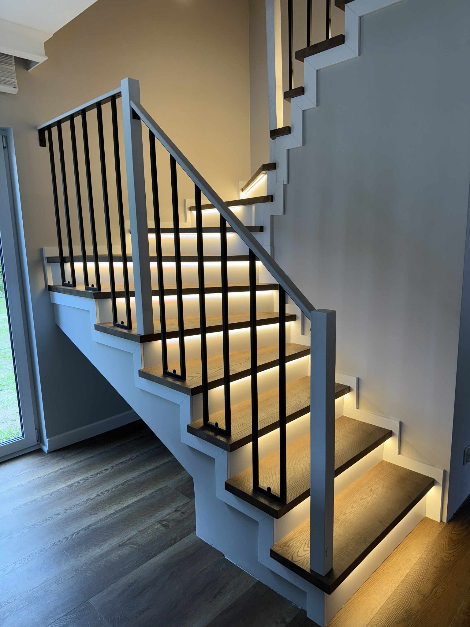 Nowoczesne schody z drewnianymi stopniami, białymi bokami i czarną balustradą. Podświetlenie LED pod stopniami tworzy unikalny efekt wizualny. Widok z dołu.