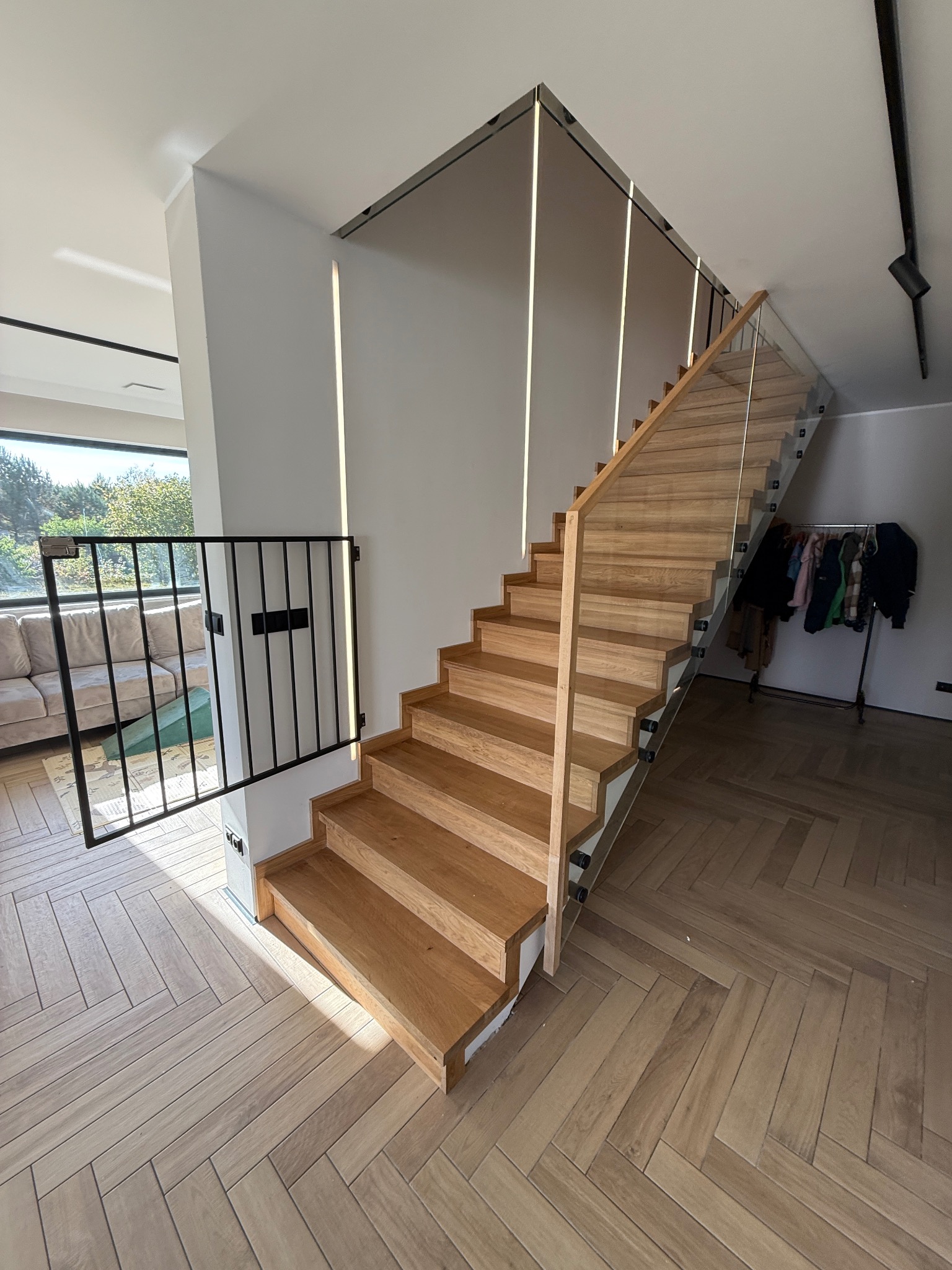 Nowoczesne schody drewniane z szklaną balustradą i zabezpieczeniem dla dzieci. Parkiet ułożony w jodełkę. Minimalistyczny design, jasne wnętrze z naturalnym światłem.