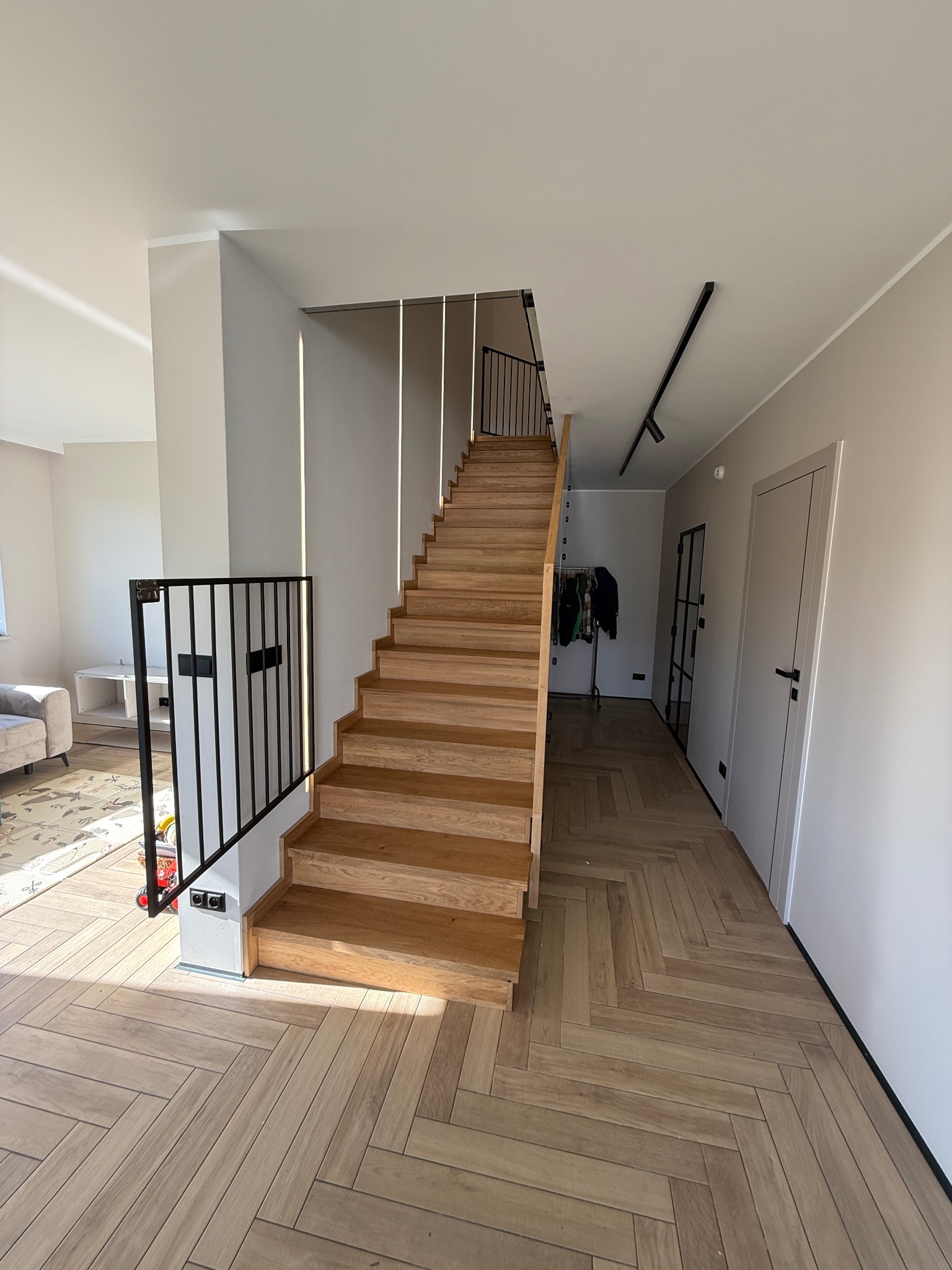 Jasne, drewniane schody z czarną, metalową balustradą w nowoczesnym wnętrzu. Parkiet ułożony w jodełkę. Minimalistyczny design, subtelne oświetlenie.