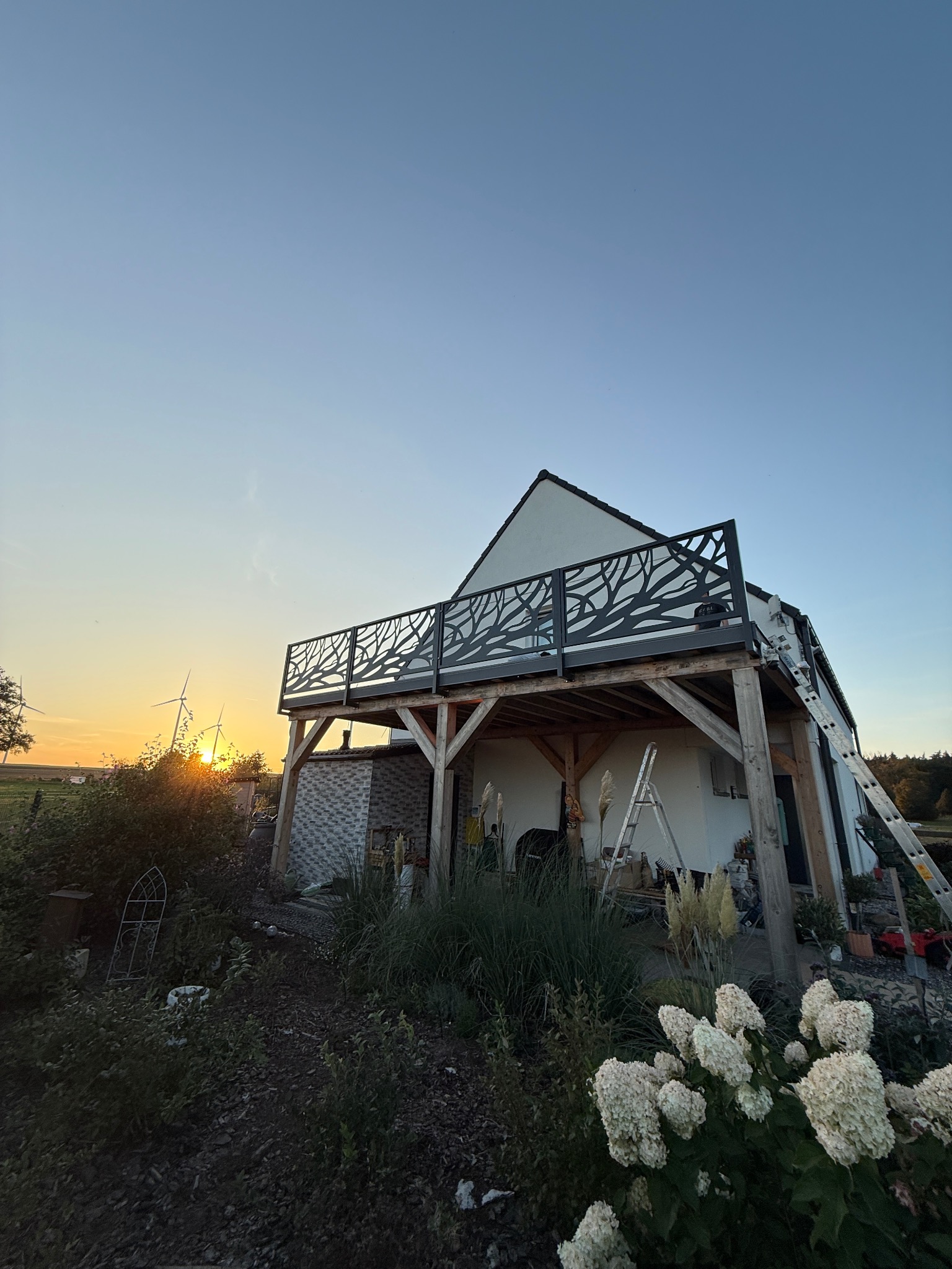 Dom z tarasem i balustradą o wzorze drzewa na tle zachodzącego słońca. Widoczne drabiny sugerują prace wykończeniowe. W tle turbiny wiatrowe.