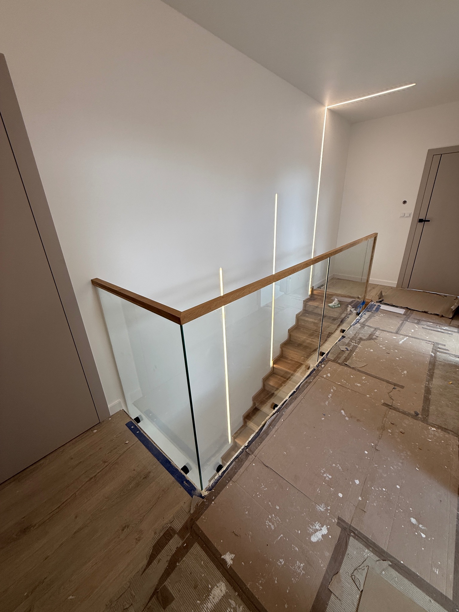 Nowoczesne schody z drewnianymi stopniami i szklaną balustradą z drewnianą poręczą. Oświetlenie LED wzdłuż ściany. Widoczne zabezpieczenia podłogi podczas montażu.