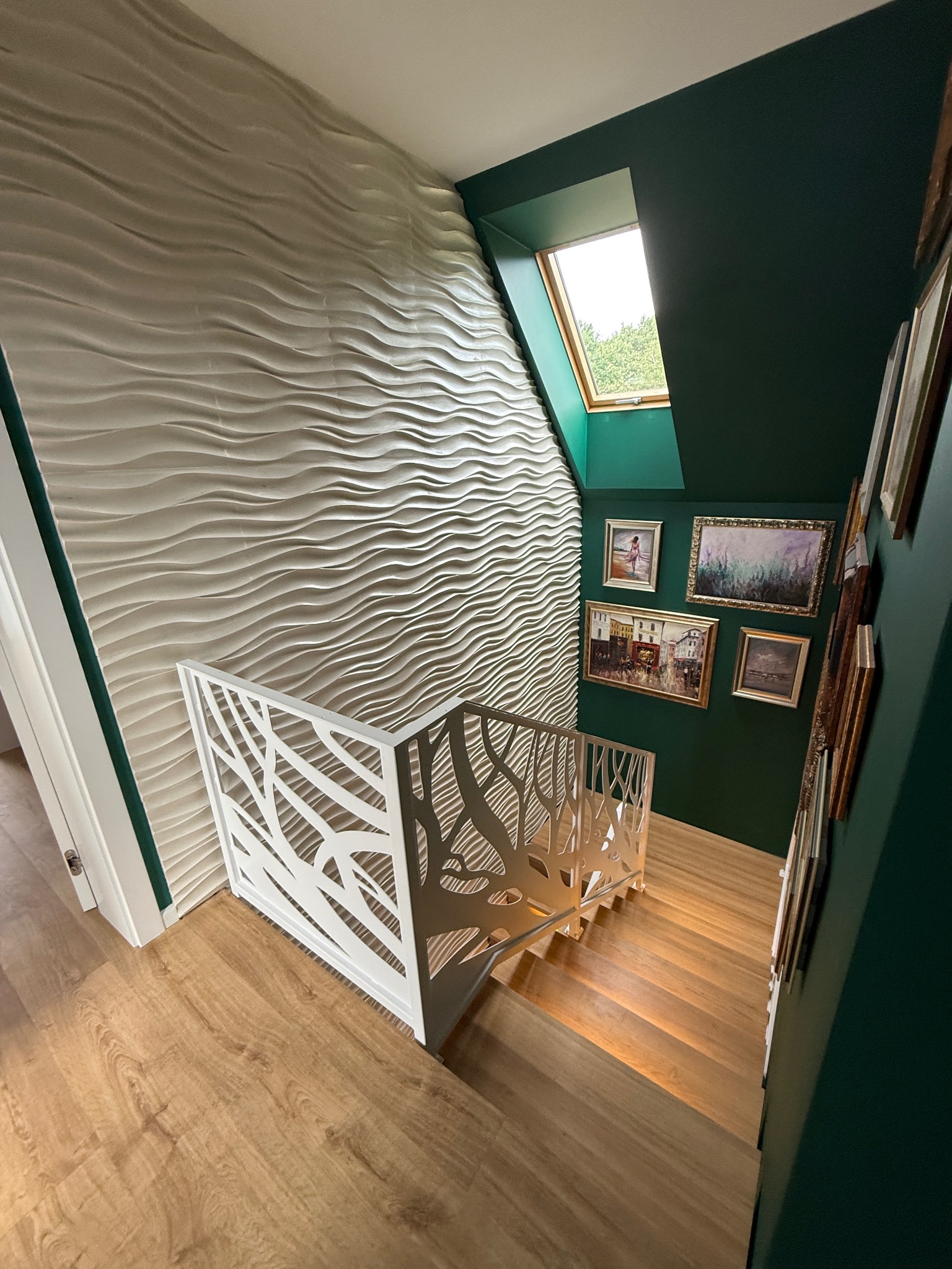 Nowoczesne schody z jasnego drewna z białą, ażurową balustradą, oświetlone od góry oknem dachowym. Ściana z dekoracyjną fakturą i galeria obrazów w ciemnozielonym wnętrzu.