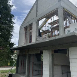 Surowy stan dwukondygnacyjnego domu z betonu kom&oacute;rkowego w trakcie budowy, widoczne rusztowania, podpory i częściowo wzniesione ściany działowe, na pierwszym planie taczka i resztki materiał&oacute;w...