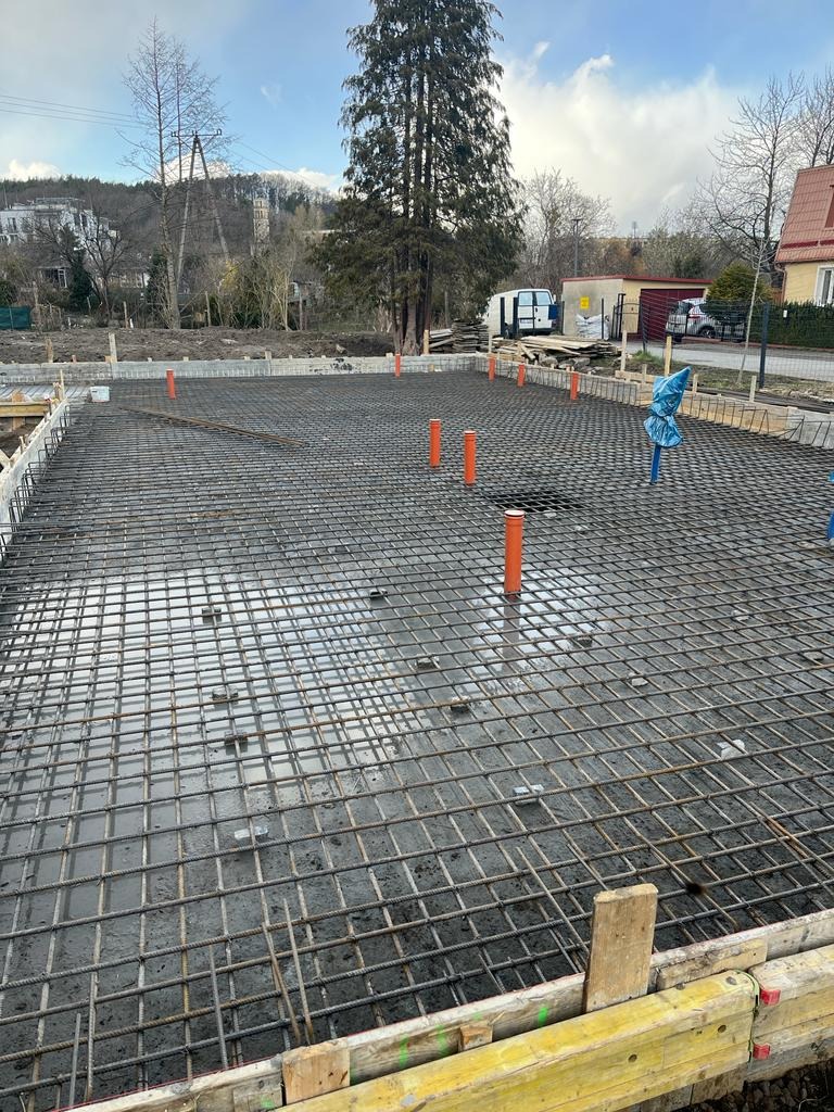 Zalany betonem szkielet zbrojeniowy fundamentu pod budynek jednorodzinny, widoczne pionowe rury kanalizacyjne i szalunki drewniane, w tle zabudowa jednorodzinna i drzewa.