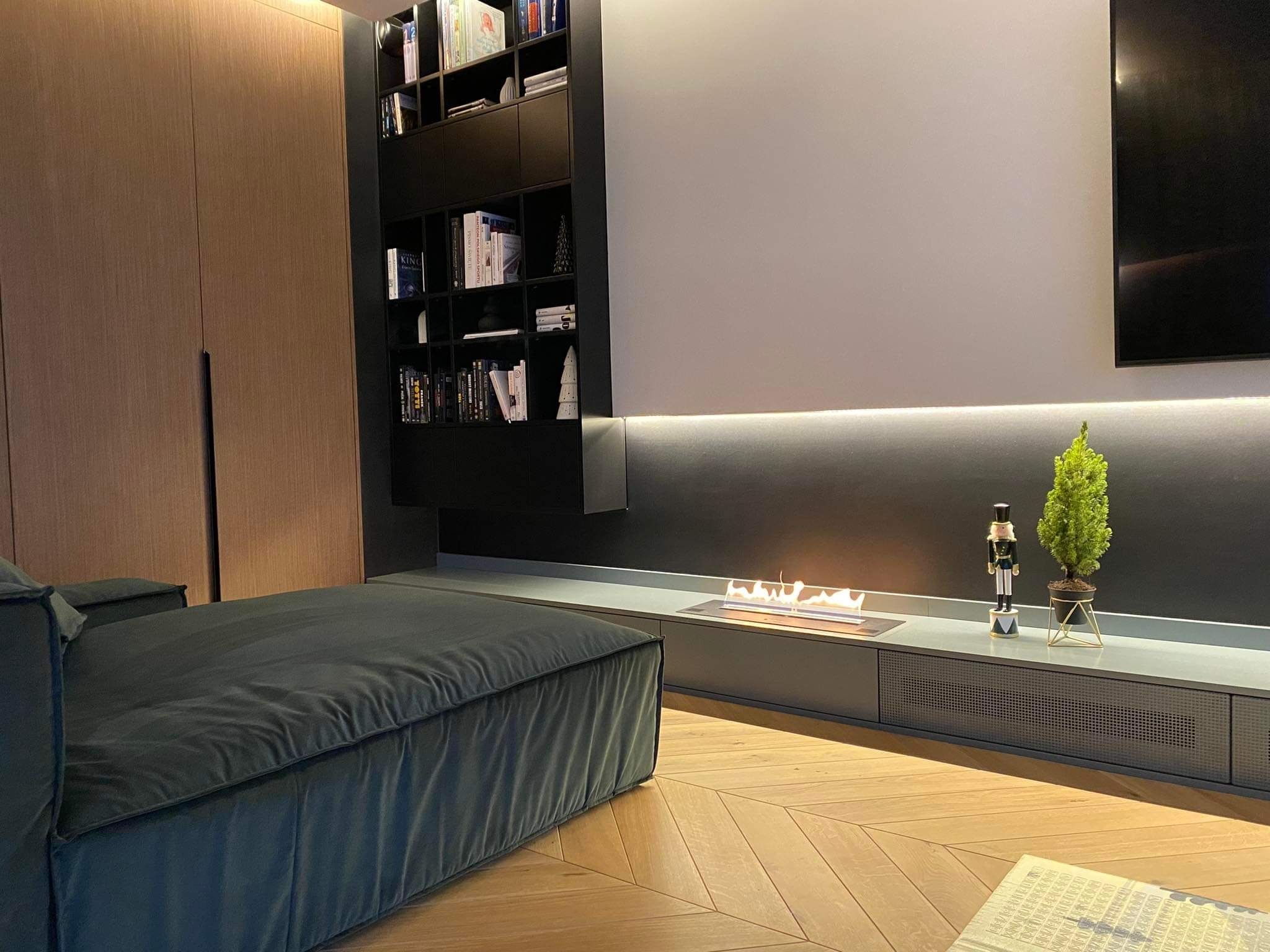 Nowoczesny salon z bio-kominkiem, drewnianą podłogą w jodełkę, ciemnoszarą zabudową meblową, czarnymi półkami na książki i zielonym narożnikiem.