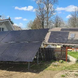 Instalacja fotowoltaiczna naziemna z widocznymi panelami na konstrukcji wsporczej oraz panelami na dachu budynku w tle, w słoneczny dzień.