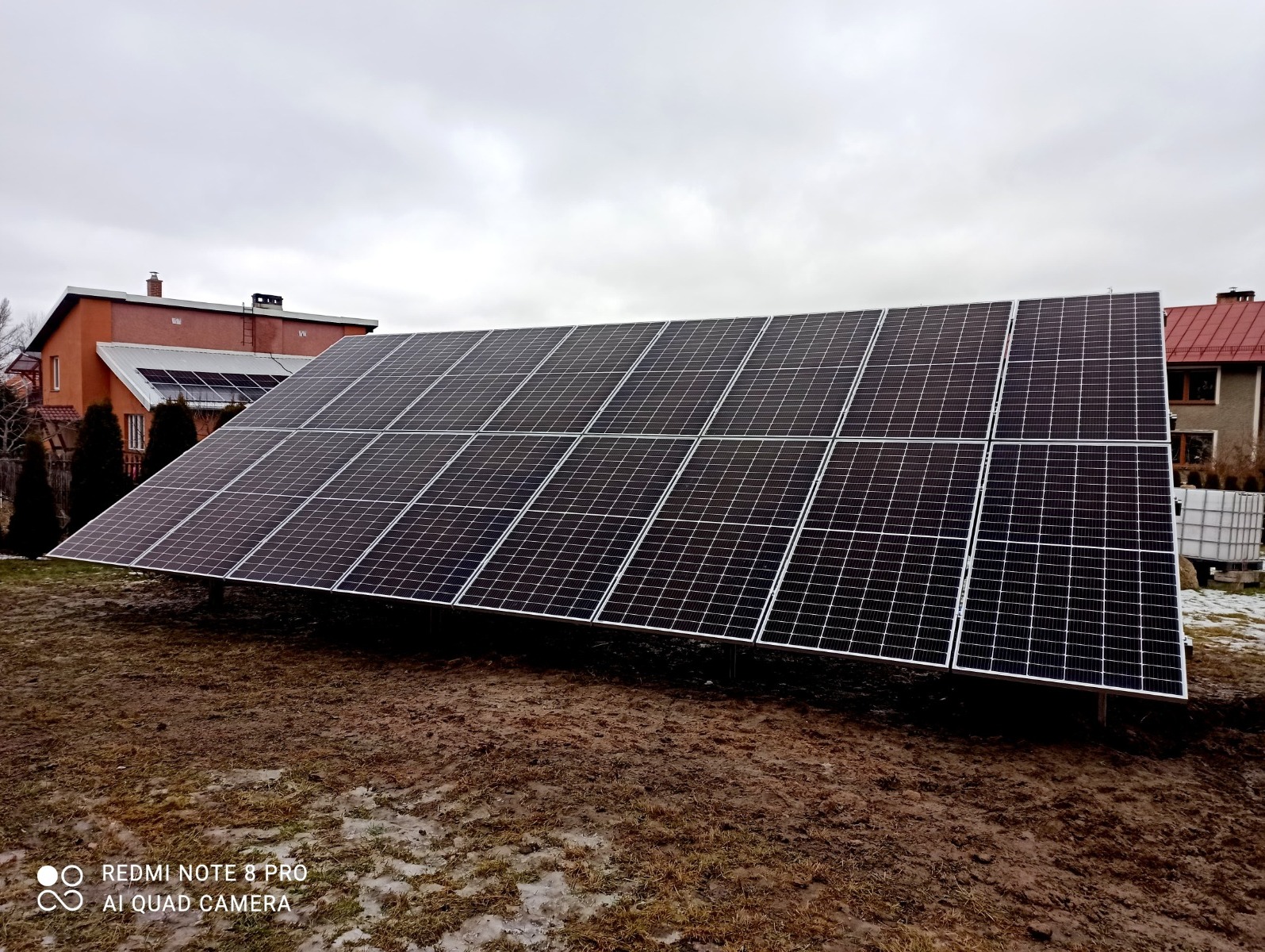 Instalacja paneli słonecznych na gruncie przed domem jednorodzinnym w pochmurny dzień. Widoczne panele na stelażu i dom w tle.