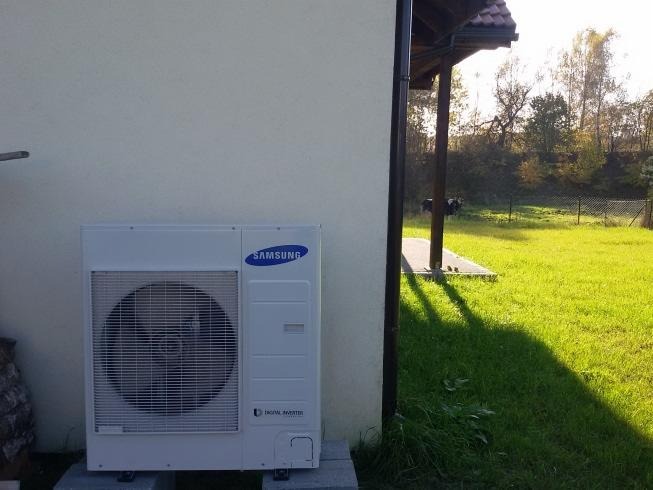 Zewnętrzna jednostka klimatyzacji Samsung zamontowana na betonowych bloczkach przy ścianie budynku z widokiem na trawnik i drzewa w tle.