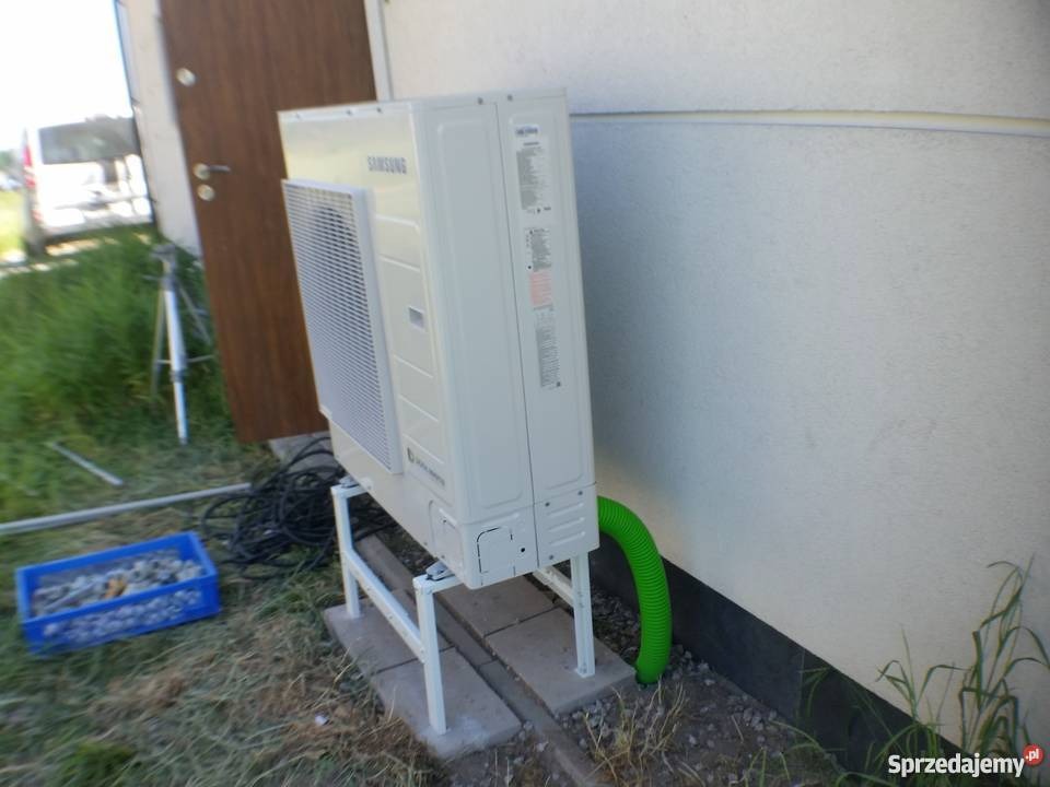 Zewnętrzna jednostka pompy ciepła Samsung na białym stelażu, z zielonym przewodem odprowadzającym wodę, ustawiona przy jasnej ścianie budynku. W tle widoczne drzwi i częściowo samochód.