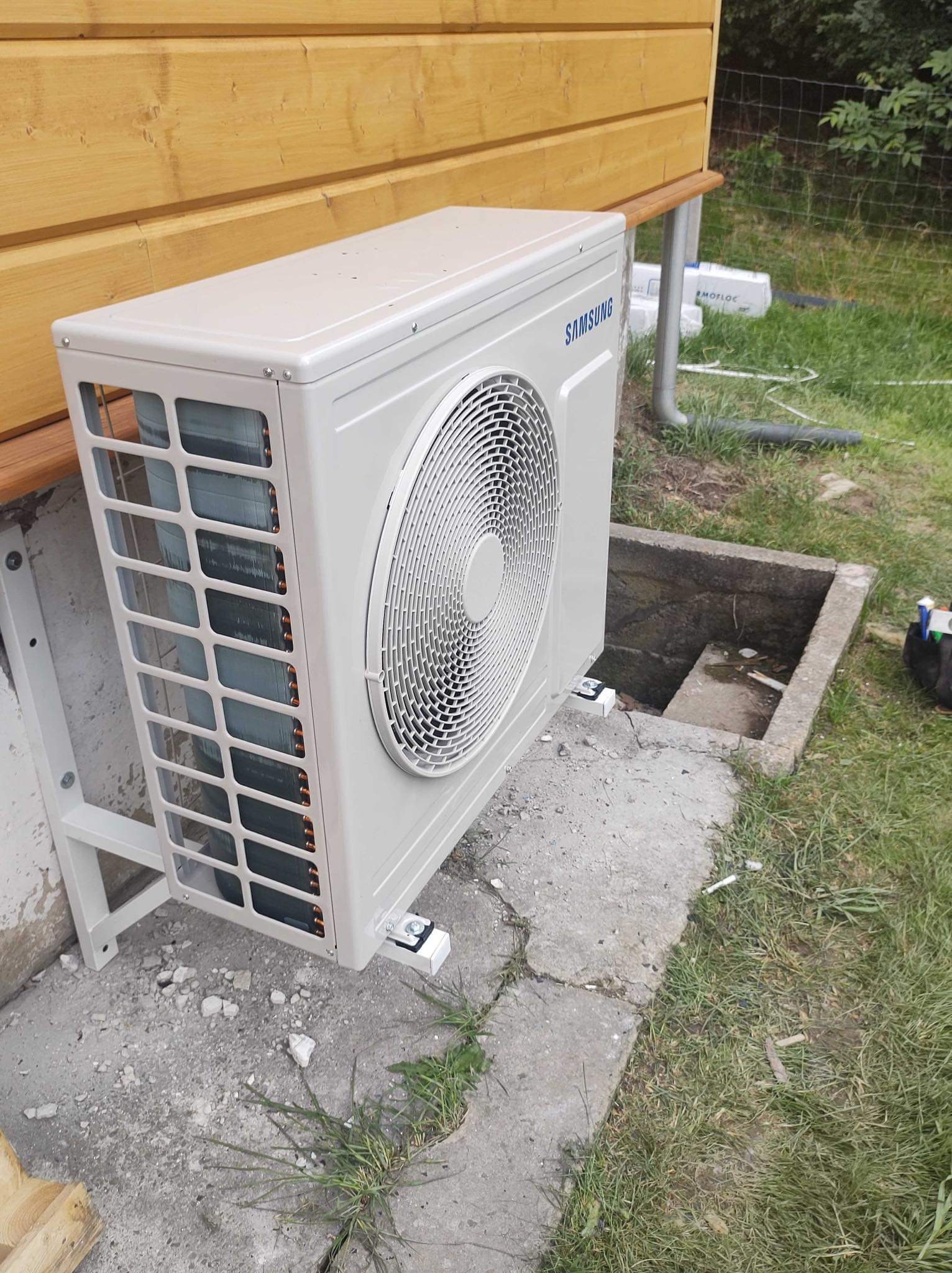Zewnętrzna jednostka klimatyzacyjna marki Samsung zamontowana na betonowym fundamencie przy drewnianej elewacji budynku, widoczne elementy instalacji i fragment trawnika.