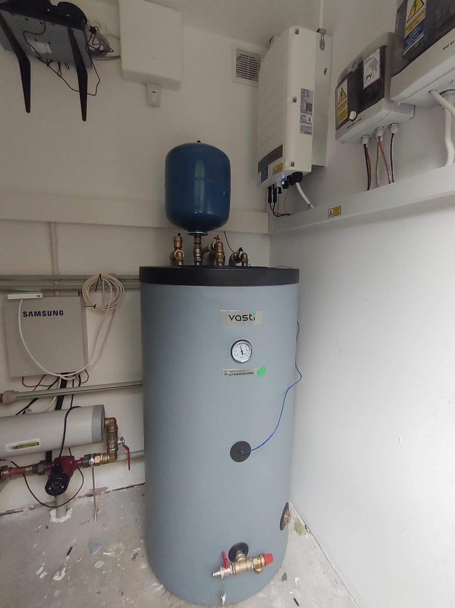 Szary zasobnik ciepłej wody użytkowej marki Vasti z niebieskim naczyniem wzbiorczym, widoczne przyłącza hydrauliczne, manometr i osprzęt elektryczny, na ścianie inwerter fotowoltaiczny i skrzynka...