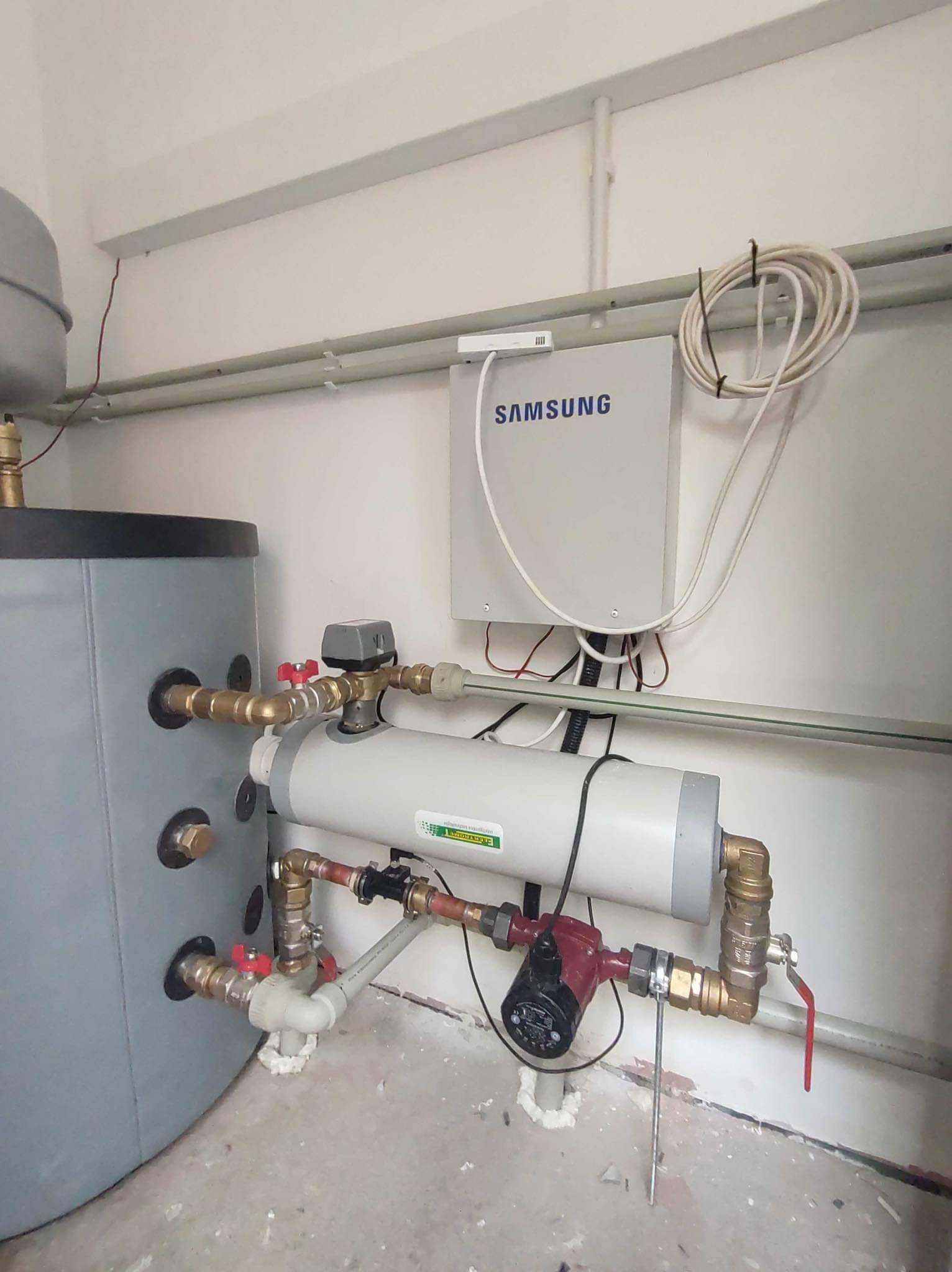 Szara instalacja pompy ciepła z zbiornikiem, pompą obiegową i sterownikiem Samsung na białej ścianie, widoczne liczne rury i zawory.