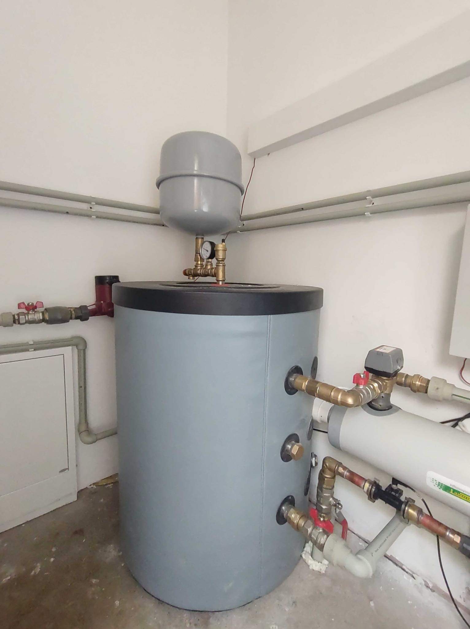 Szary akumulator ciepła w kotłowni z widocznymi rurami i zaworami, podłączony do systemu grzewczego, z manometrem na górze zbiornika.