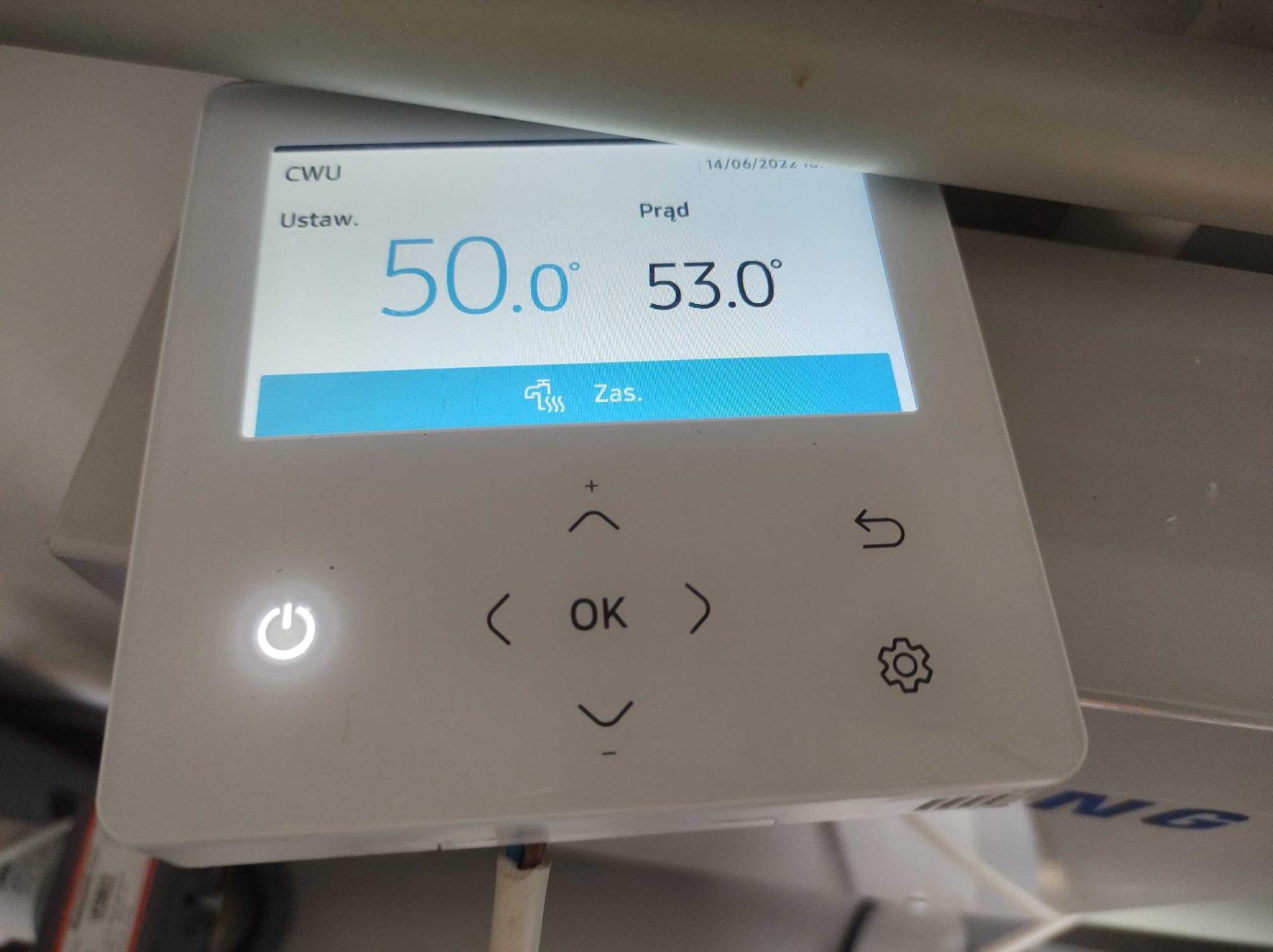 Panel sterowania pompy ciepła z wyświetlaną temperaturą wody użytkowej ustawioną na 50 stopni Celsjusza i aktualną temperaturą 53 stopni Celsjusza. Widoczne przyciski nawigacyjne i symbol zasilania.