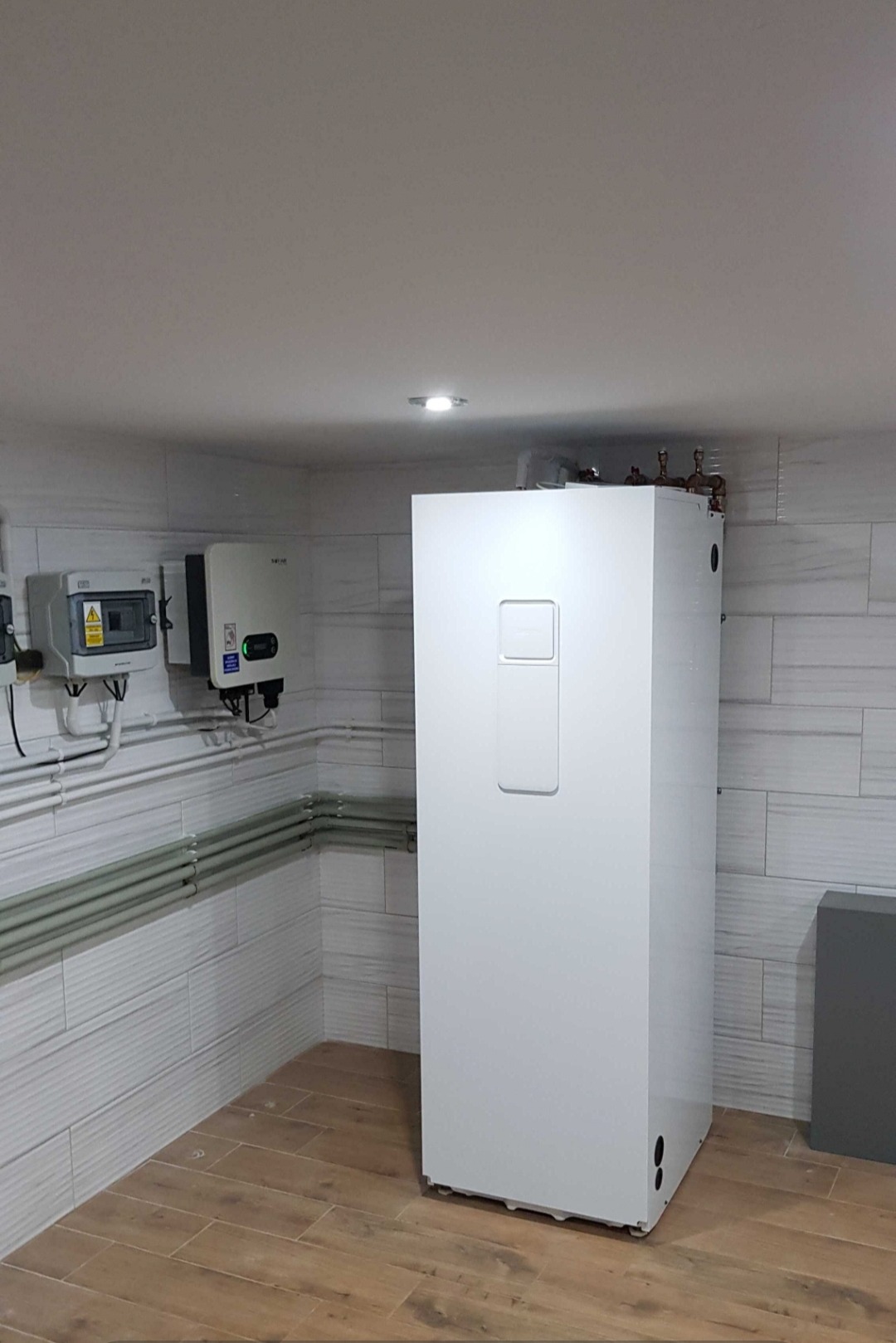 Biała, prostopadłościenna jednostka w pomieszczeniu z panelami elektrycznymi i rurami na ścianie, drewnopodobna podłoga.