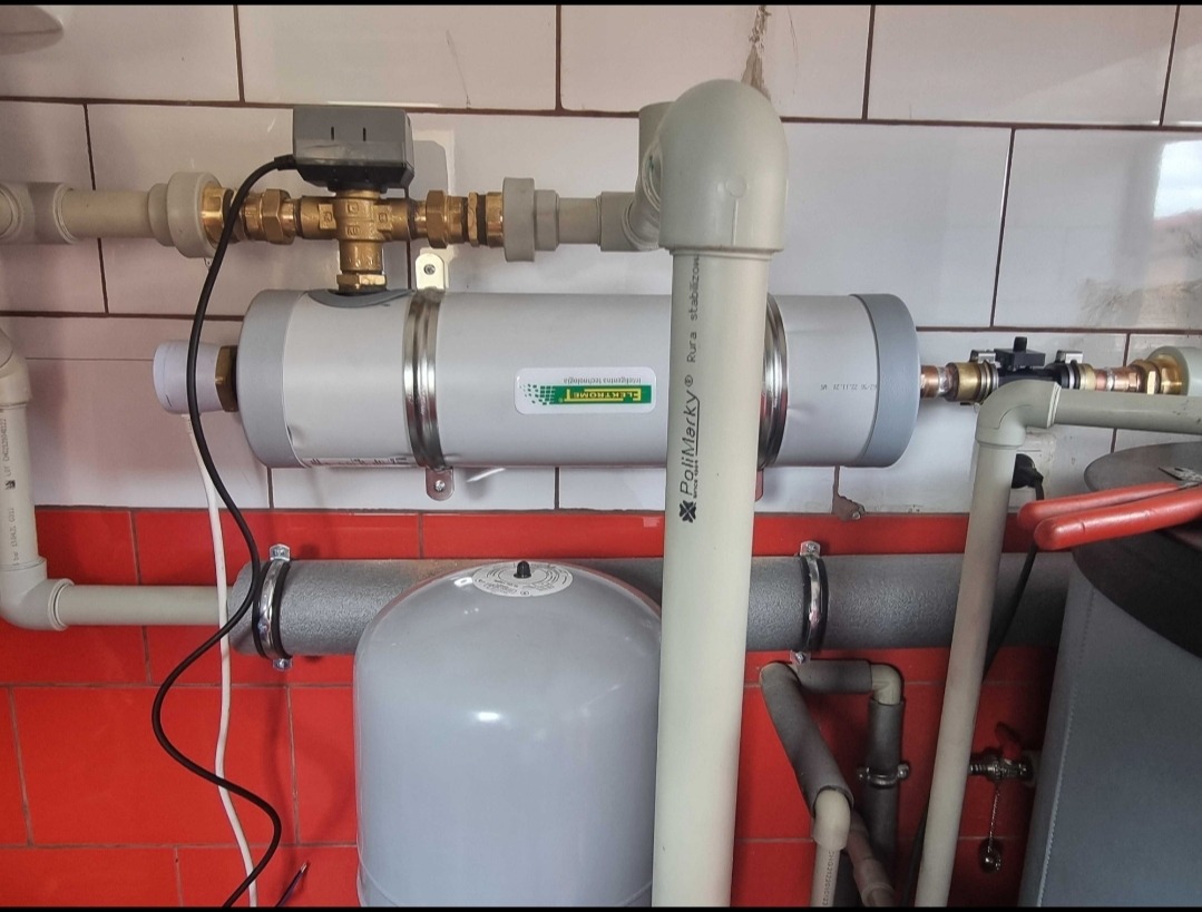 Zamontowany poziomo szary filtr wody z zieloną etykietą i rurami, widoczne rury PCV, zawory i zbiornik wyrównawczy na tle czerwono-białych płytek.