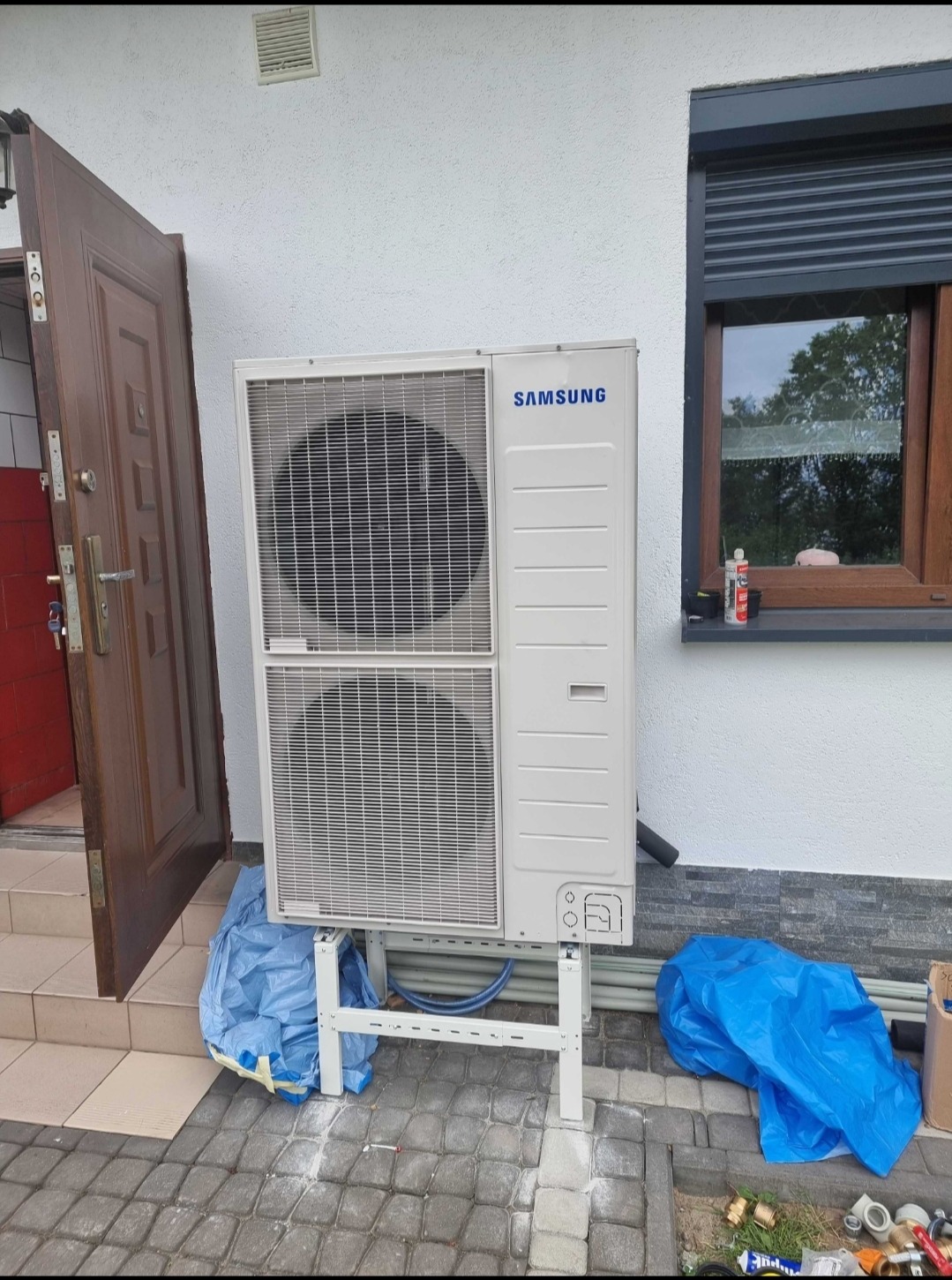 Zewnętrzna jednostka klimatyzacji Samsung na białym stelażu, obok otwartych brązowych drzwi wejściowych i okna z roletą, zabezpieczona folią budowlaną, widoczne rury i akcesoria instalacyjne