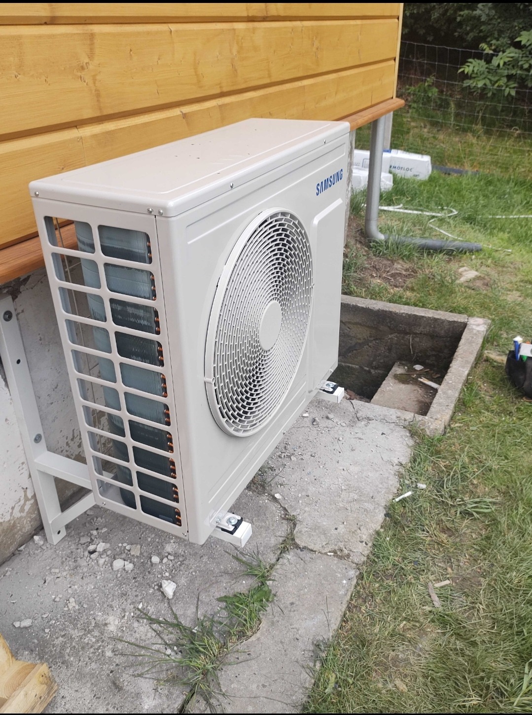 Zewnętrzna jednostka klimatyzacji Samsung zamontowana na betonowym fundamencie przy drewnianej ścianie budynku, widoczne elementy montażowe i trawa.