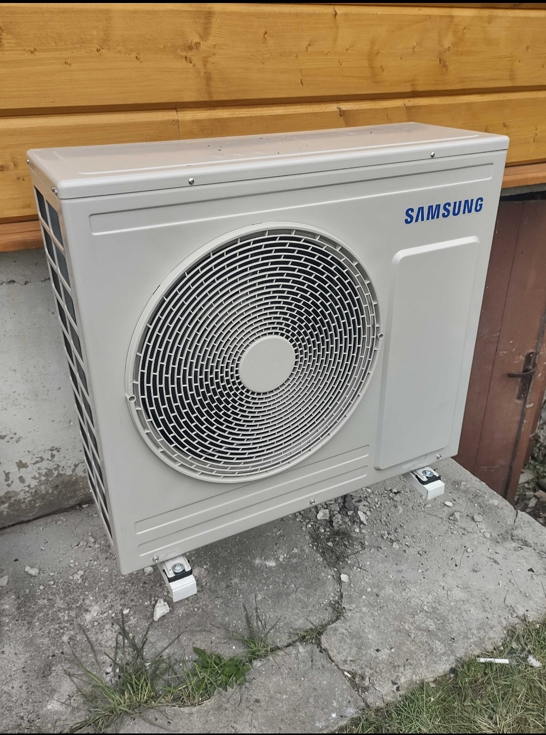 Zewnętrzna jednostka klimatyzacji marki Samsung zamontowana na betonowej podstawie przy drewnianej ścianie budynku.
