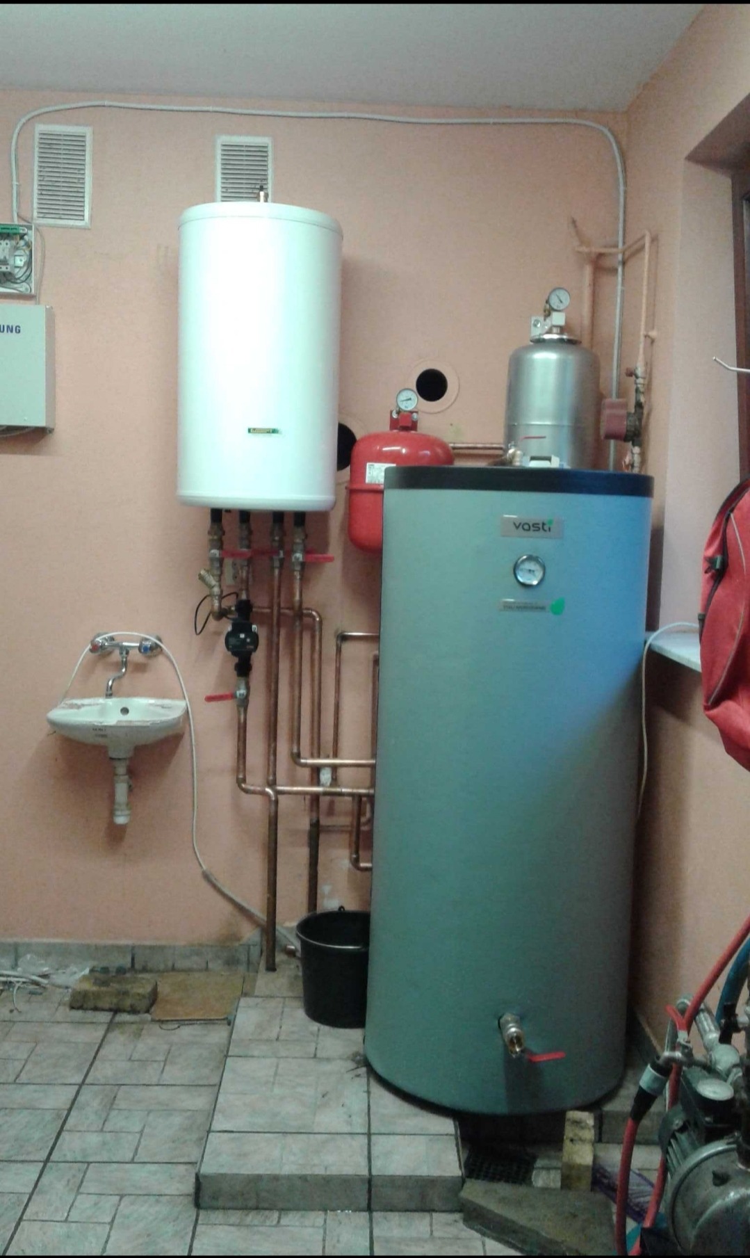 Dwa zbiorniki akumulacyjne ciepłej wody użytkowej, biały i szary, z widocznymi rurami miedzianymi, zaworami, manometrami i instalacją elektryczną na tle różowej ściany i ceramicznej podłogi...