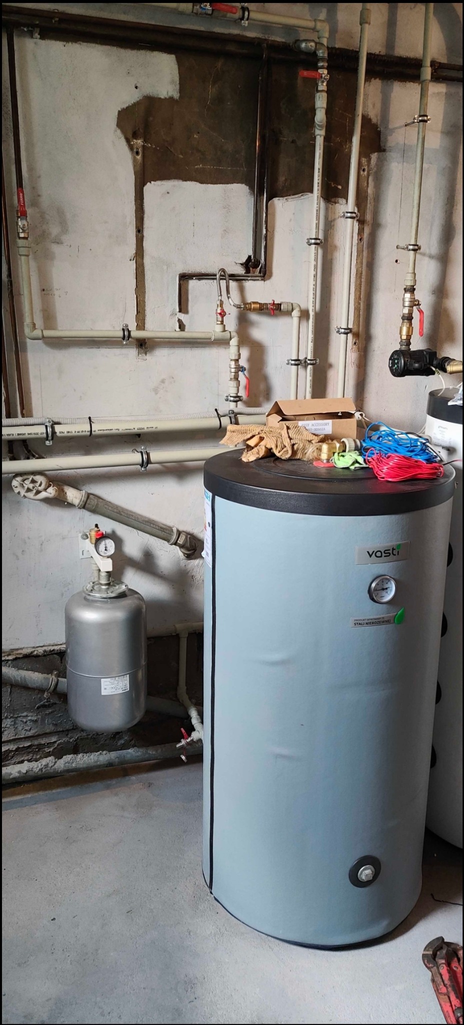 Szary zbiornik buforowy Vosti w piwnicy z widocznymi rurami, zaworami, manometrem i akcesoriami montażowymi na górze. W tle surowe ściany.