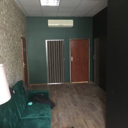 RoBo Usługi Budowlane - Wnętrze po remoncie: zielona ściana, brązowe drzwi, sufit kasetonowy, podłoga z płytek imitujących drewno, zielona sofa z rzeczami na niej.