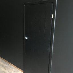 RoBo Usługi Budowlane - Ciemne drzwi z metalową klamką, zamontowane w czarnej ścianie, widok na fragment podłogi z płytek imitujących drewno.