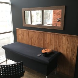 RoBo Usługi Budowlane - Pokój z łóżkiem do masażu przykrytym czarnym materiałem, drewnianym zagłówkiem i lustrem, w którym odbija się ceglana ściana i lampa pierścieniowa.