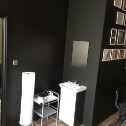 RoBo Usługi Budowlane - Czarne ściany w salonie kosmetycznym z umywalką, lustrem, lampą podłogową i wózkiem na kosmetyki, zdjęcia w ramkach na ścianie.