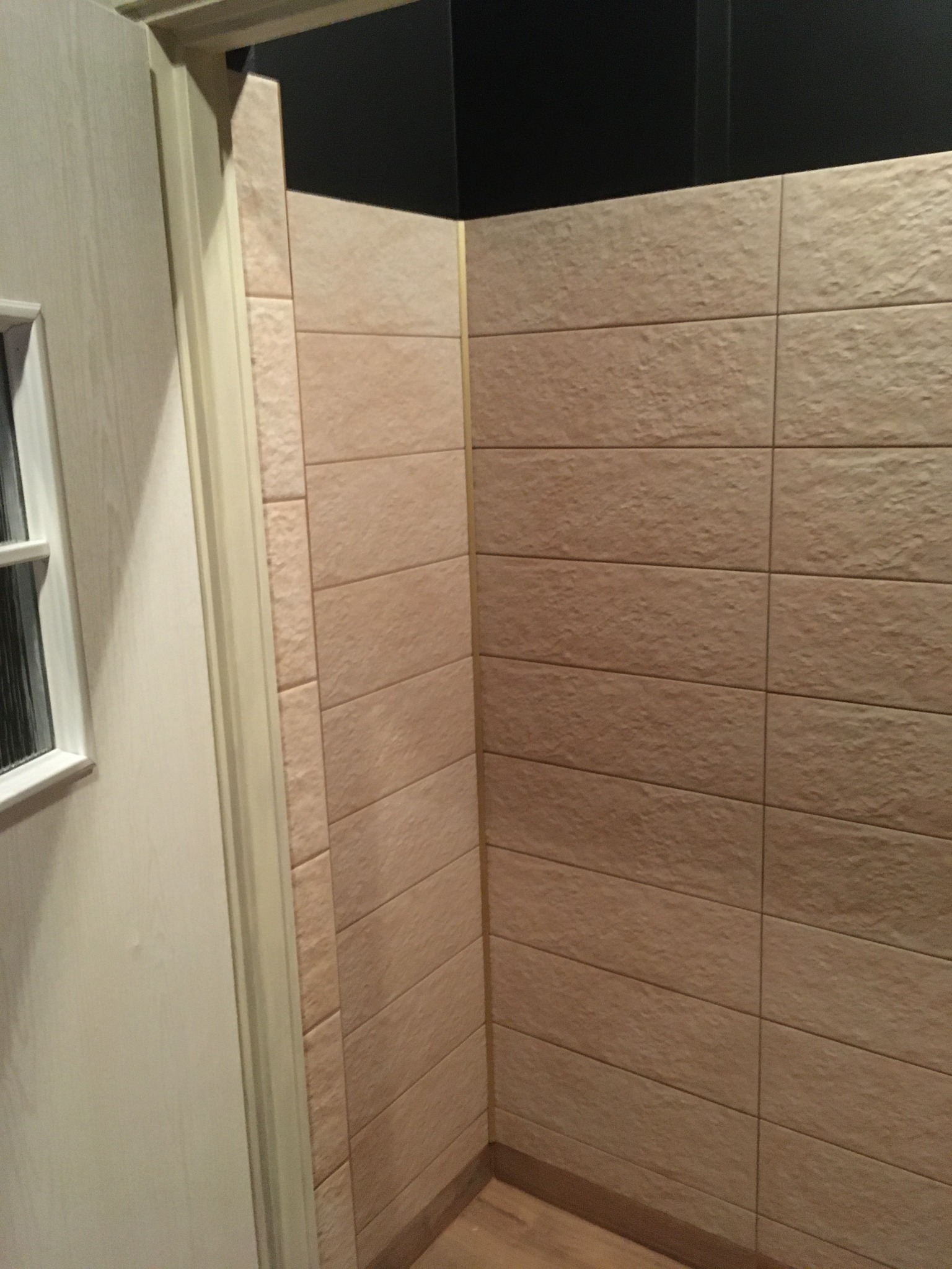 Beżowe, poziome płytki imitujące cegłę na ścianie łazienki, widoczne przy futrynie drzwi z oknem.