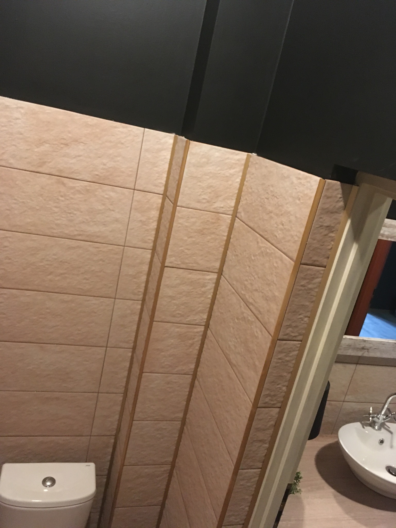 Wykończona łazienka z beżowymi płytkami ściennymi, ciemnym sufitem, białą umywalką i toaletą, widoczne narożniki z dekoracyjnymi listwami.