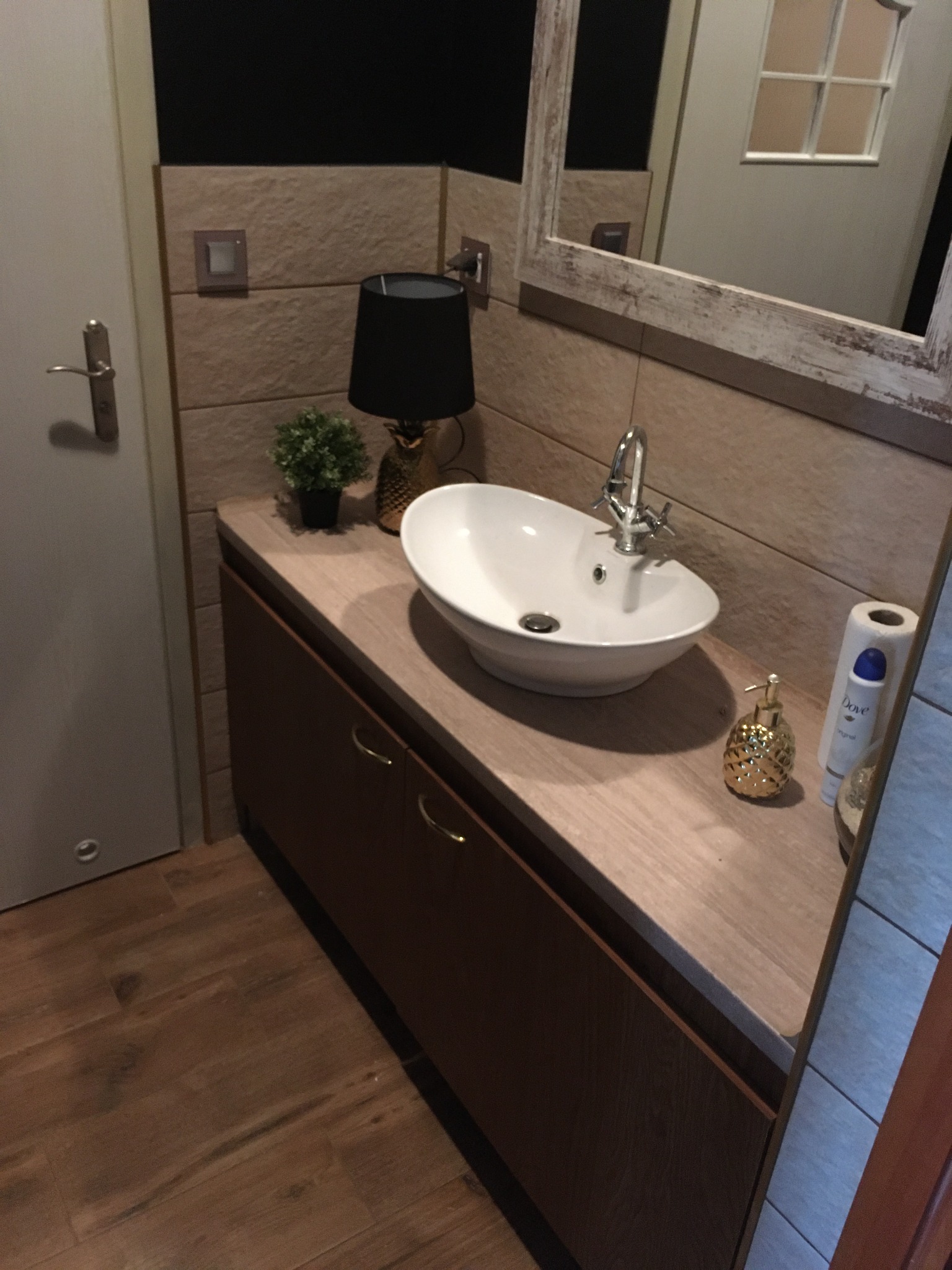 Umywalka nablatowa w kształcie łódki z chromowaną baterią, ustawiona na szafce z ciemnego drewna, dekorowana lampką w kształcie ananasa i małą rośliną doniczkową, na tle ściany wyłożonej płytkami...