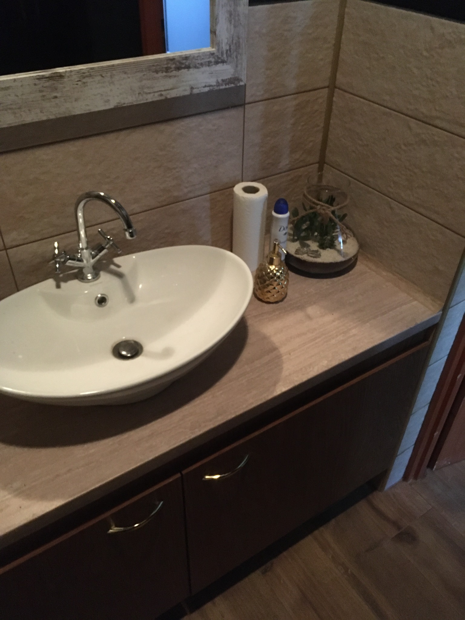 Umywalka nablatowa z chromowaną baterią, obok dozownik mydła w kształcie ananasa, rolka papieru toaletowego, kosmetyk i dekoracyjny terrarium w szklanym naczyniu na blacie z drewnopodobną okleiną...