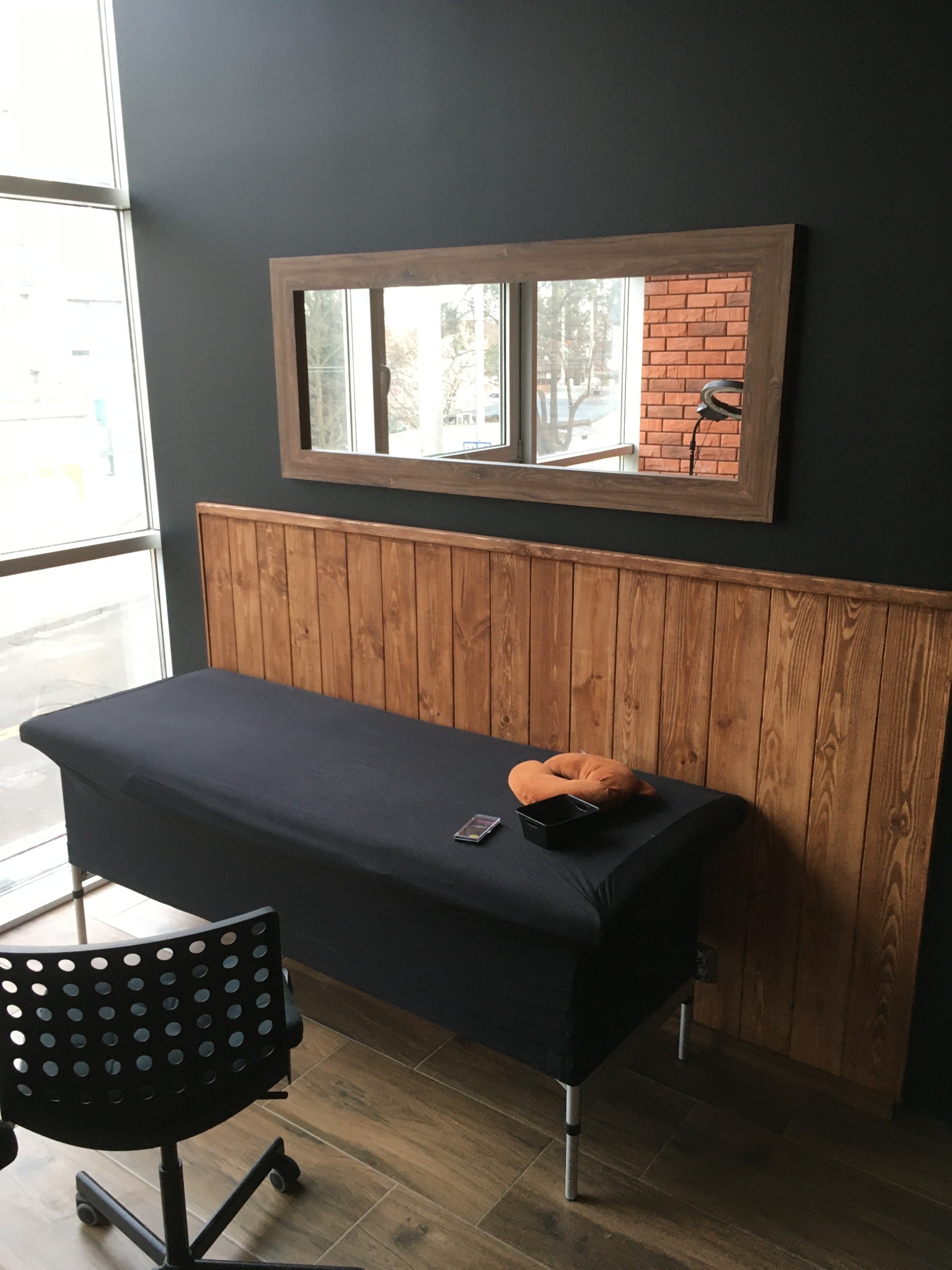 Pokój z łóżkiem do masażu przykrytym czarnym materiałem, drewnianym zagłówkiem i lustrem, w którym odbija się ceglana ściana i lampa pierścieniowa.