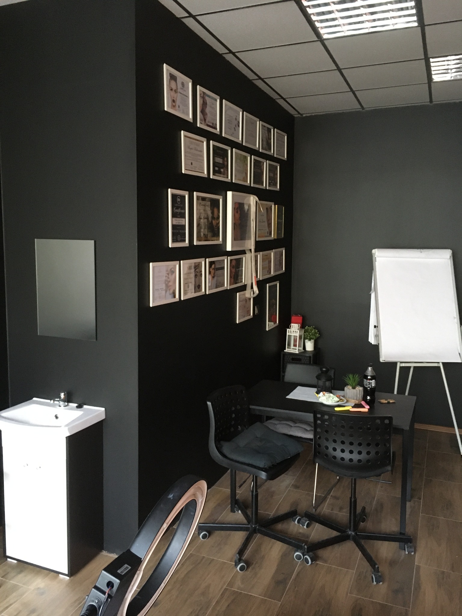 Wnętrze gabinetu kosmetycznego z czarnymi ścianami, umywalką, lustrem, stołem z krzesłami na kółkach, tablicą suchościeralną oraz ścianą ozdobioną ramkami z certyfikatami i zdjęciami.