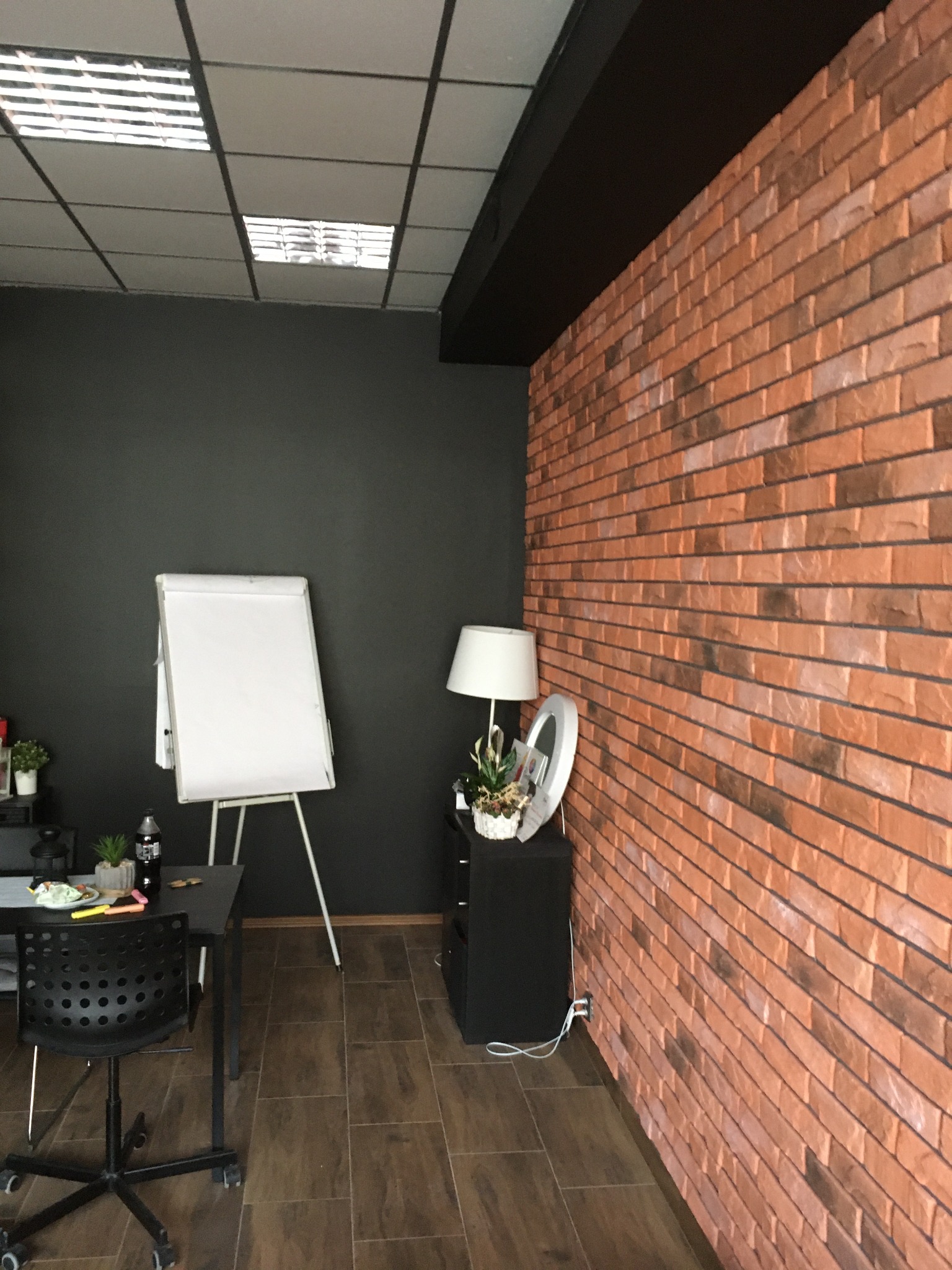 Biuro z wykończoną ścianą z cegły dekoracyjnej w odcieniach brązu, czarną ścianą, flipchartem, biurkiem z krzesłem na kółkach i lampą.