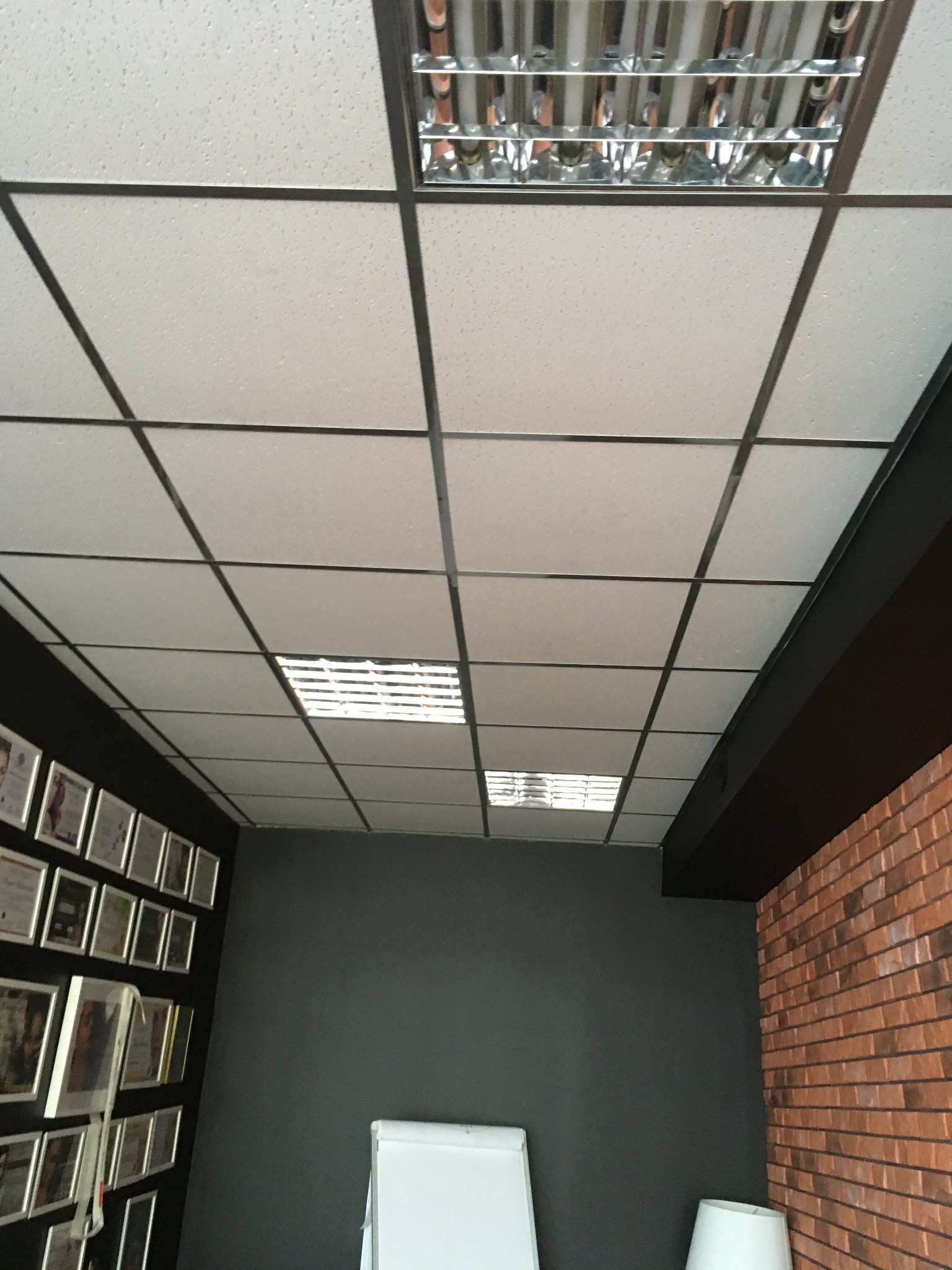 Kasetonowy sufit podwieszany z rastrowymi oprawami oświetleniowymi, widok z dołu, ściana z dekoracyjną cegłą i ściana z certyfikatami w ramkach w tle.