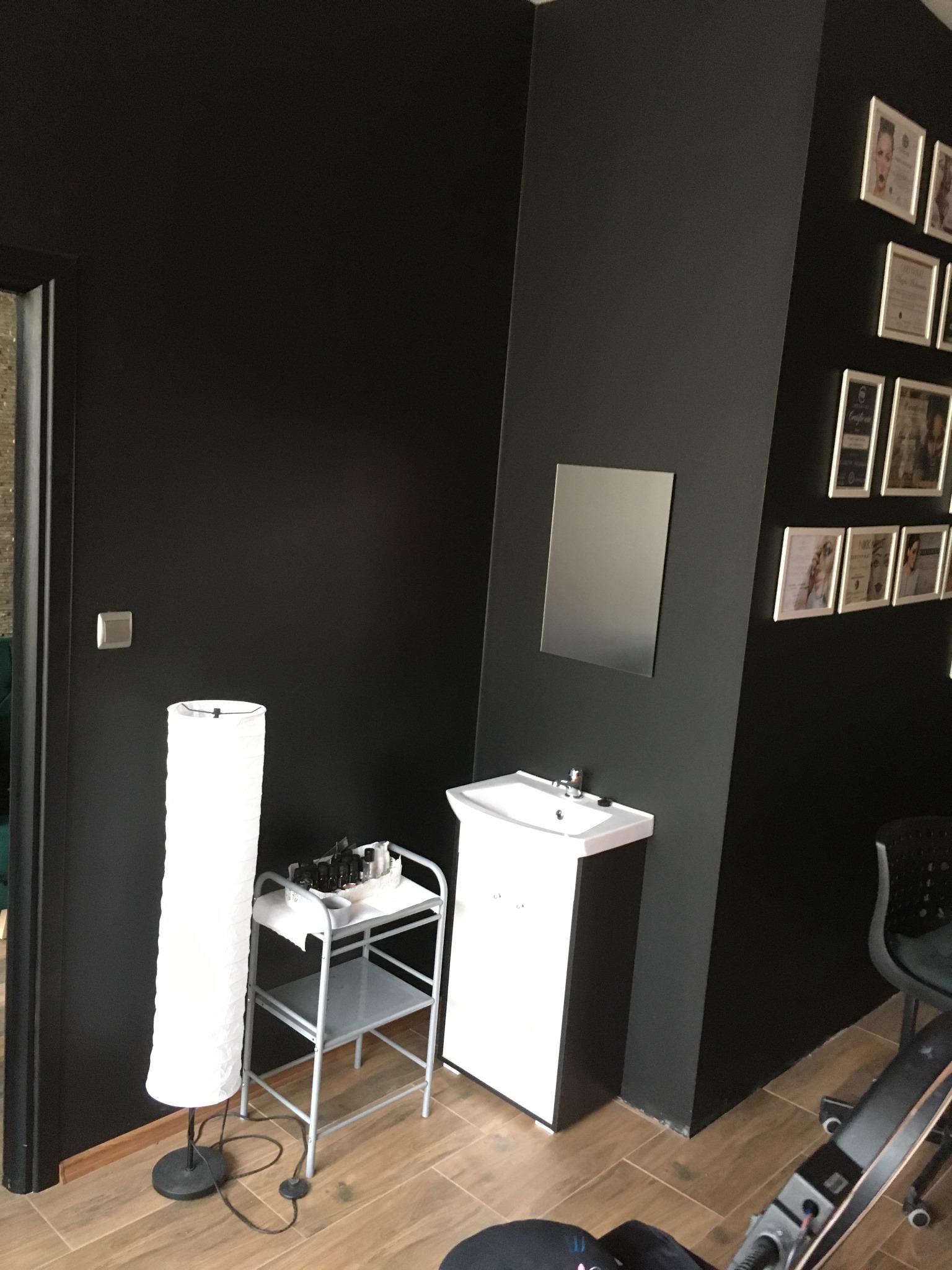 Czarne ściany w salonie kosmetycznym z umywalką, lustrem, lampą podłogową i wózkiem na kosmetyki, zdjęcia w ramkach na ścianie.