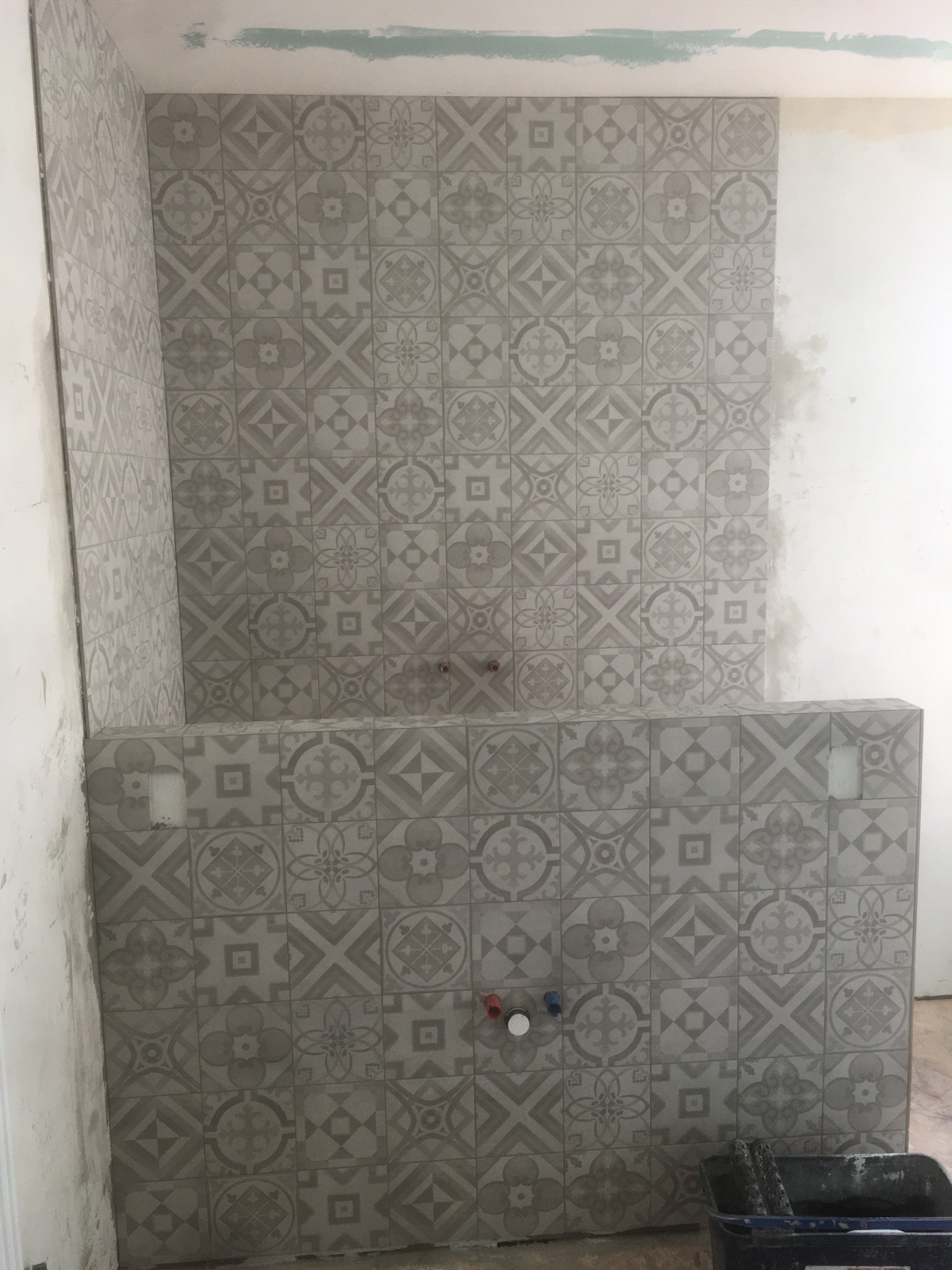 Fragment łazienki w trakcie wykańczania, ściany wyłożone szarymi płytkami ceramicznymi z motywem kwiatowym i geometrycznym, widoczne otwory na armaturę i pojemnik z narzędziami glazurniczymi.