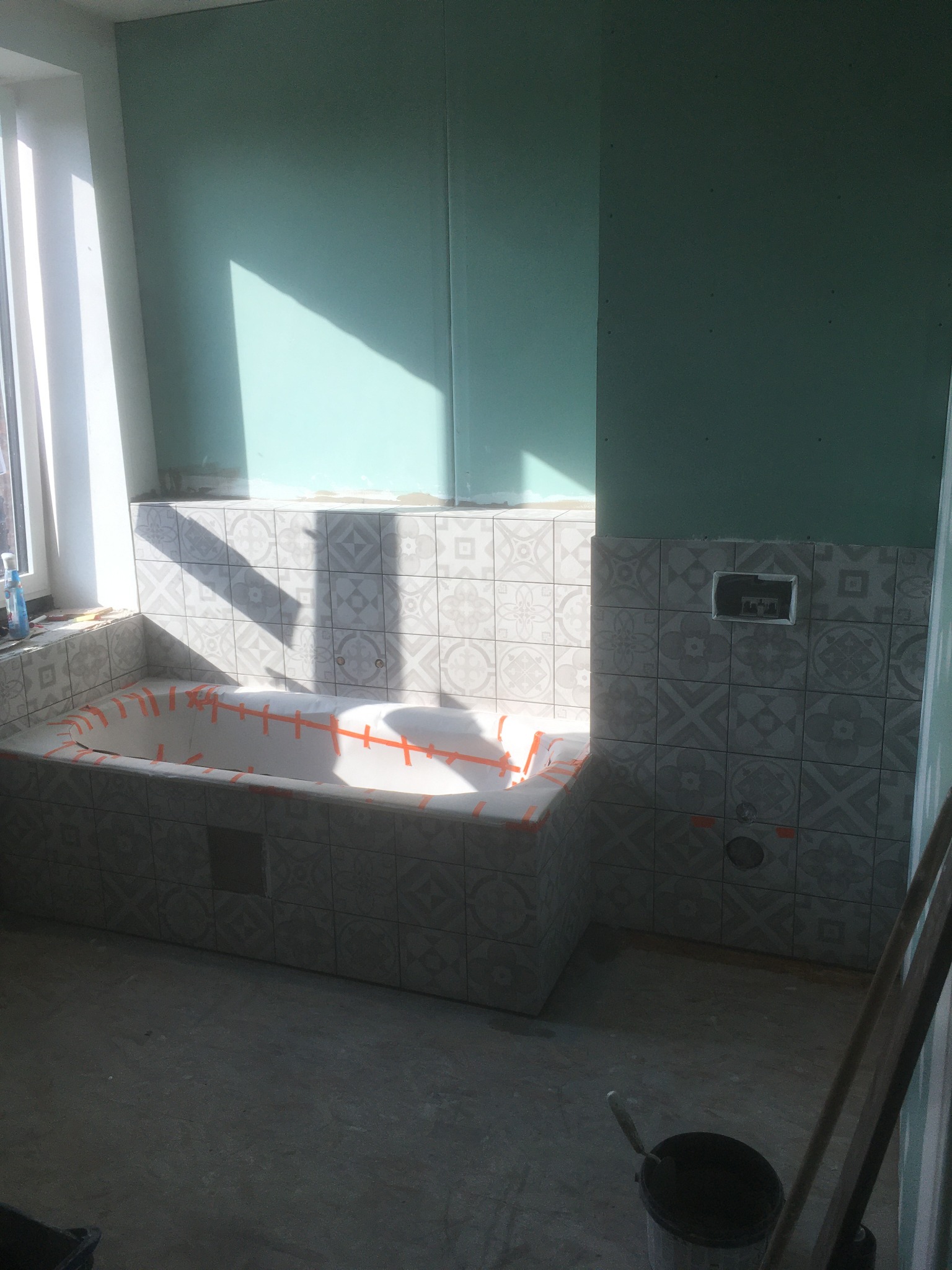 Wnętrze łazienki w trakcie remontu: obudowana wanna zabezpieczona taśmą, częściowo wyłożona wzorzystymi płytkami ceramicznymi, ściany przygotowane do dalszych prac, widoczne narzędzia i wiadro.