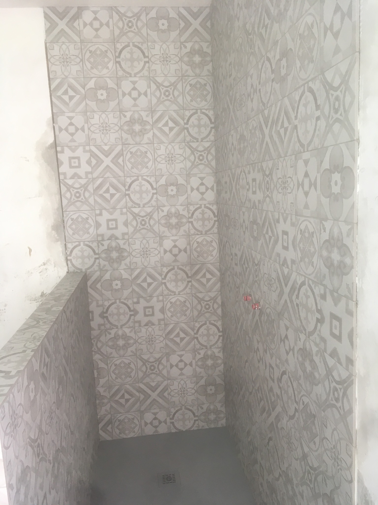 Świeżo wyłożona, wzorzysta glazura w odcieniach szarości na ścianach i podłodze w narożnym prysznicu, widoczne czerwone kliny dystansowe.