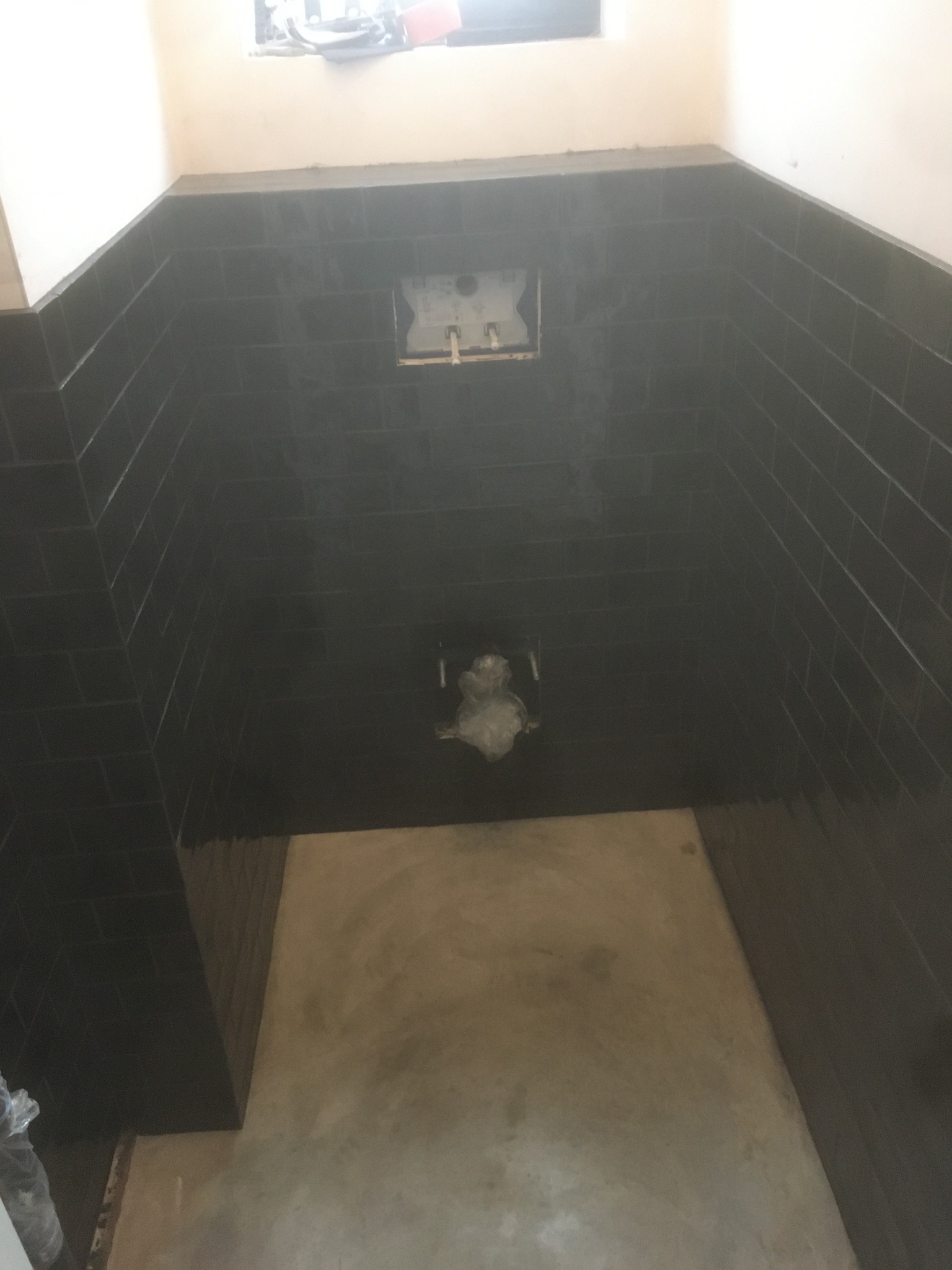 Świeżo położone, ciemne, błyszczące płytki ścienne w małej łazience w trakcie remontu, widoczne przyłącza do toalety i surowy beton na podłodze.