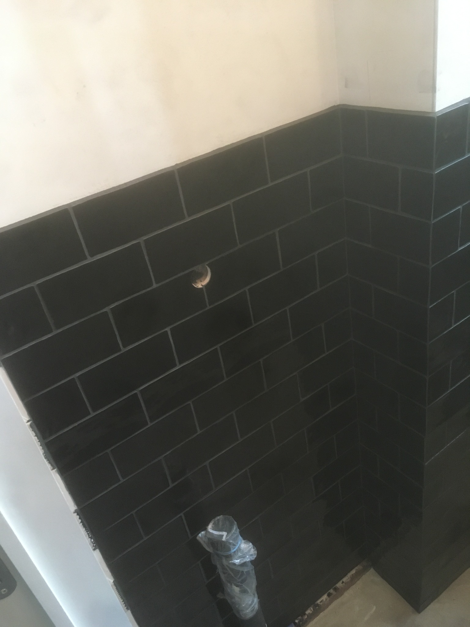 Ściana wyłożona czarnymi, prostokątnymi płytkami ceramicznymi z jasną fugą, widoczny otwór na rurę i fragment rury w folii ochronnej, dolny róg ściany z widocznym fragmentem podłogi.