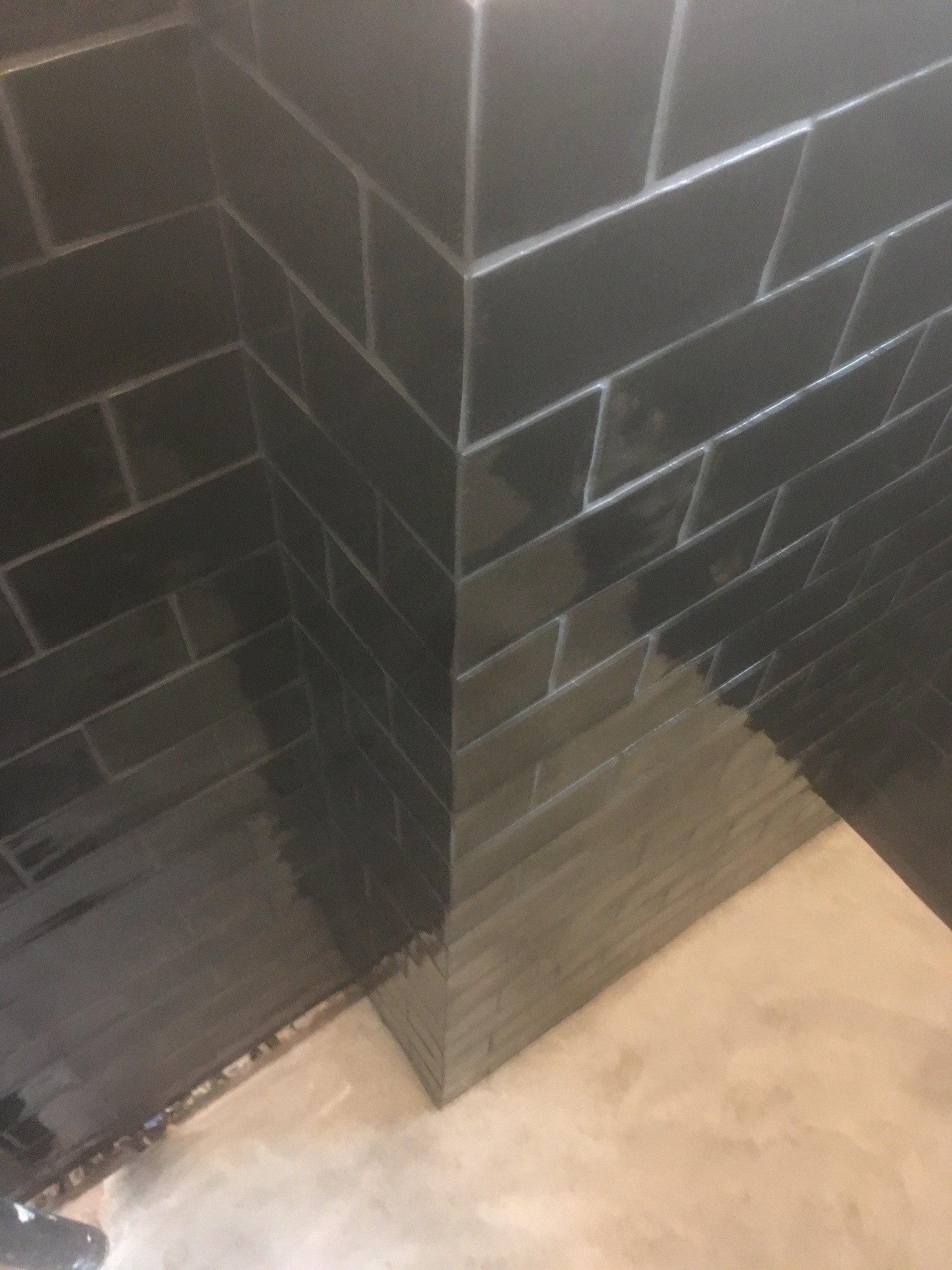 Ciemne, błyszczące płytki ułożone na ścianie w rogu pomieszczenia, widoczny fragment podłogi w trakcie prac wykończeniowych.