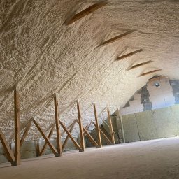 PUR-Izolacja.pl - Poddasze po ociepleniu pianką PUR, widoczne drewniane elementy konstrukcyjne i fragment ściany z pustaków. Izolacja równomiernie pokrywa skośny dach.