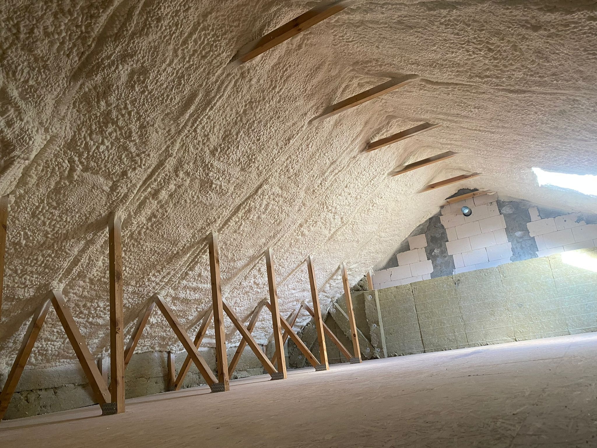Poddasze po ociepleniu pianką PUR, widoczne drewniane elementy konstrukcyjne i fragment ściany z pustaków. Izolacja równomiernie pokrywa skośny dach.