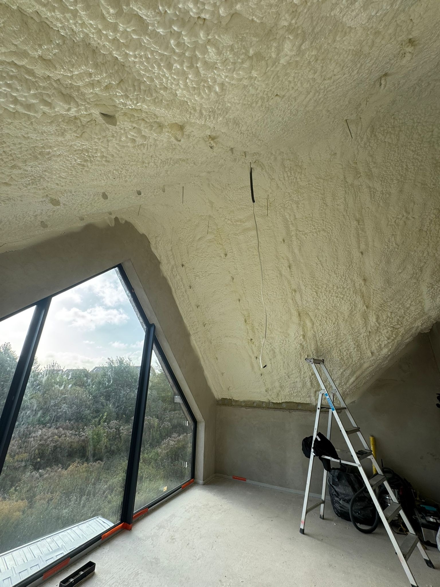 Poddasze po ociepleniu pianką PUR, widoczne okno z widokiem na zieleń, drabina oparta o ścianę. Biała, nierówna powierzchnia pianki pokrywa skosy dachu.