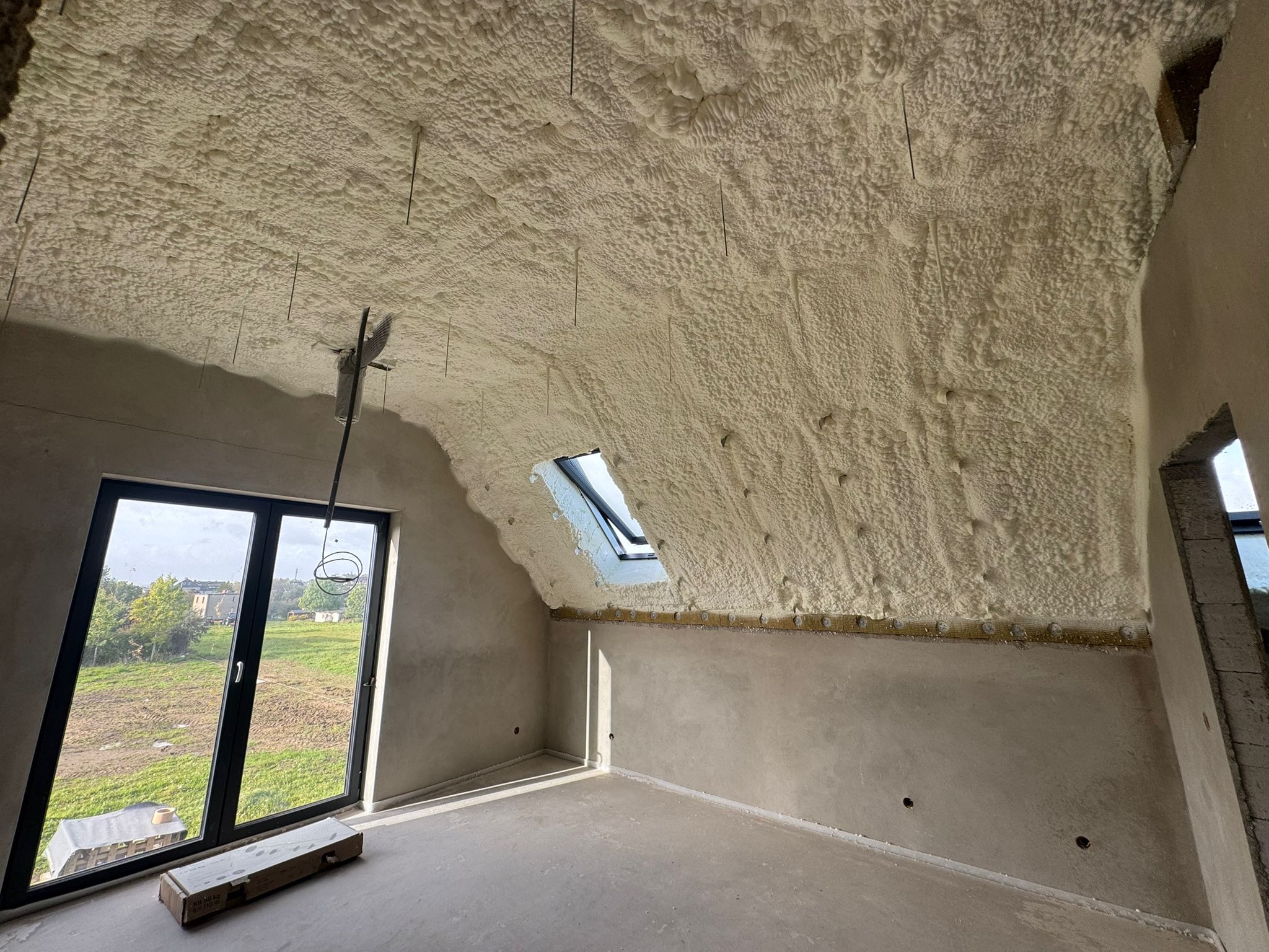 Poddasze po ociepleniu pianką PUR, widoczne okno dachowe i fragment ściany. Na podłodze karton. Widok na zewnątrz przez duże okno.