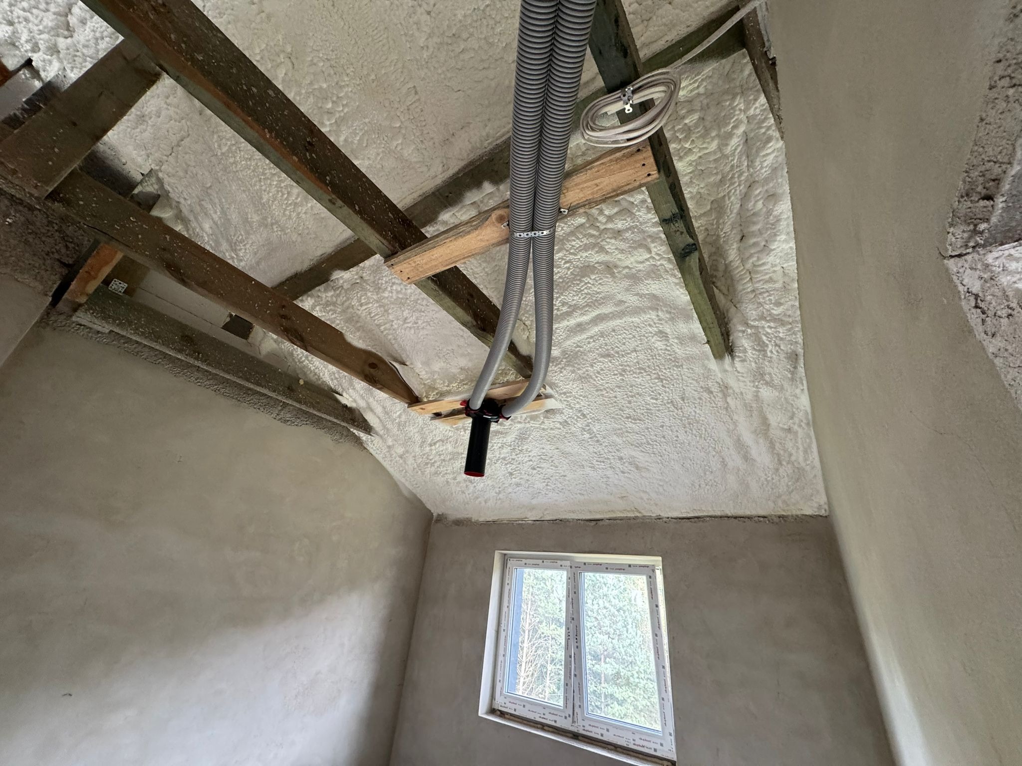 Poddasze po ociepleniu pianką PUR: widoczne drewniane belki, szara pianka, szare rury w peszlu i okno z widokiem na drzewa. Ściany w surowym tynku.