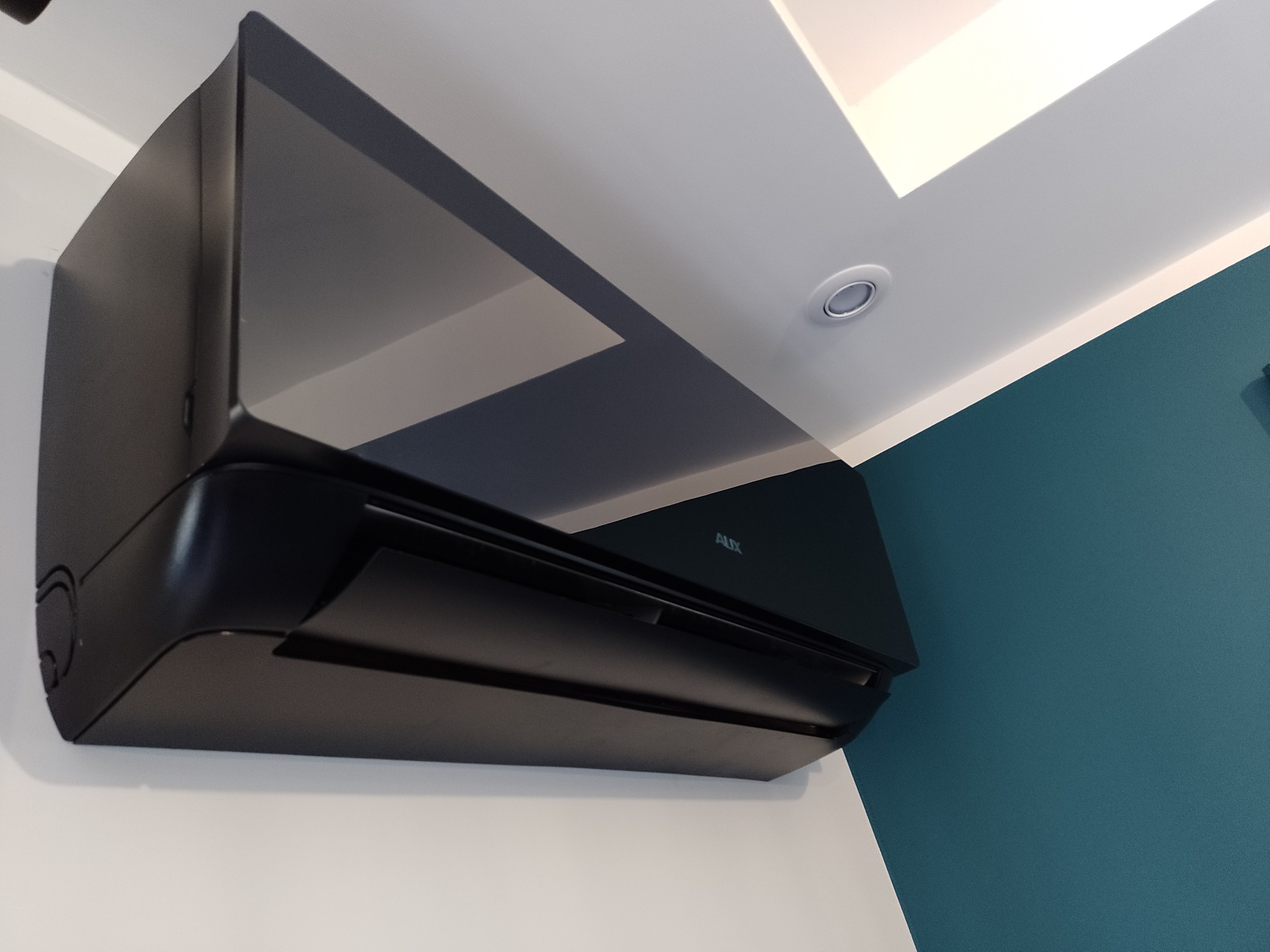 Czarna, błyszcząca jednostka wewnętrzna klimatyzacji marki AUX zamontowana na styku białej i turkusowej ściany w pomieszczeniu.